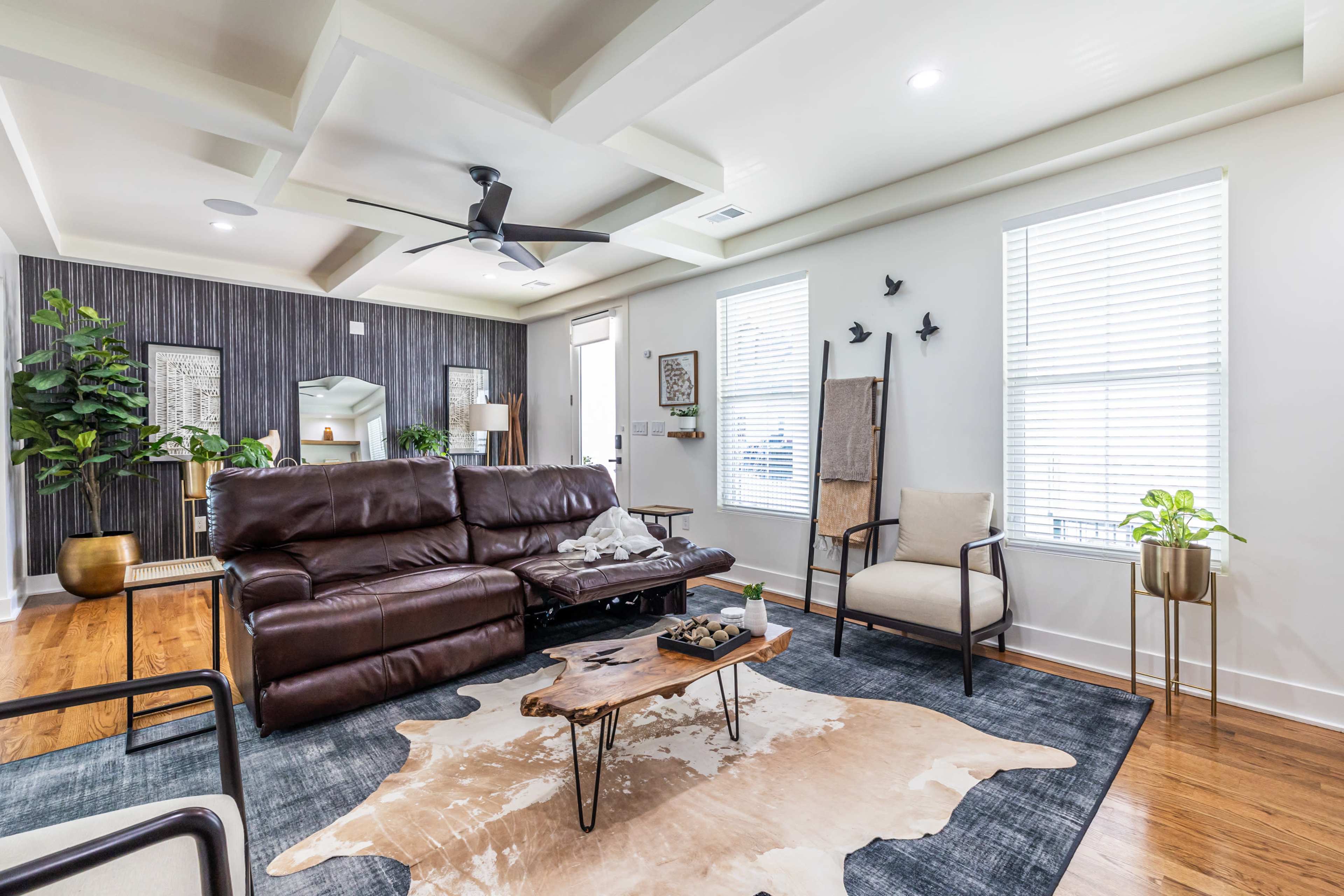 The image shows a modern living room featuring a brown leather sofa, a wooden coffee table, and two armchairs, with decorative plants and wall art enhancing the space.
