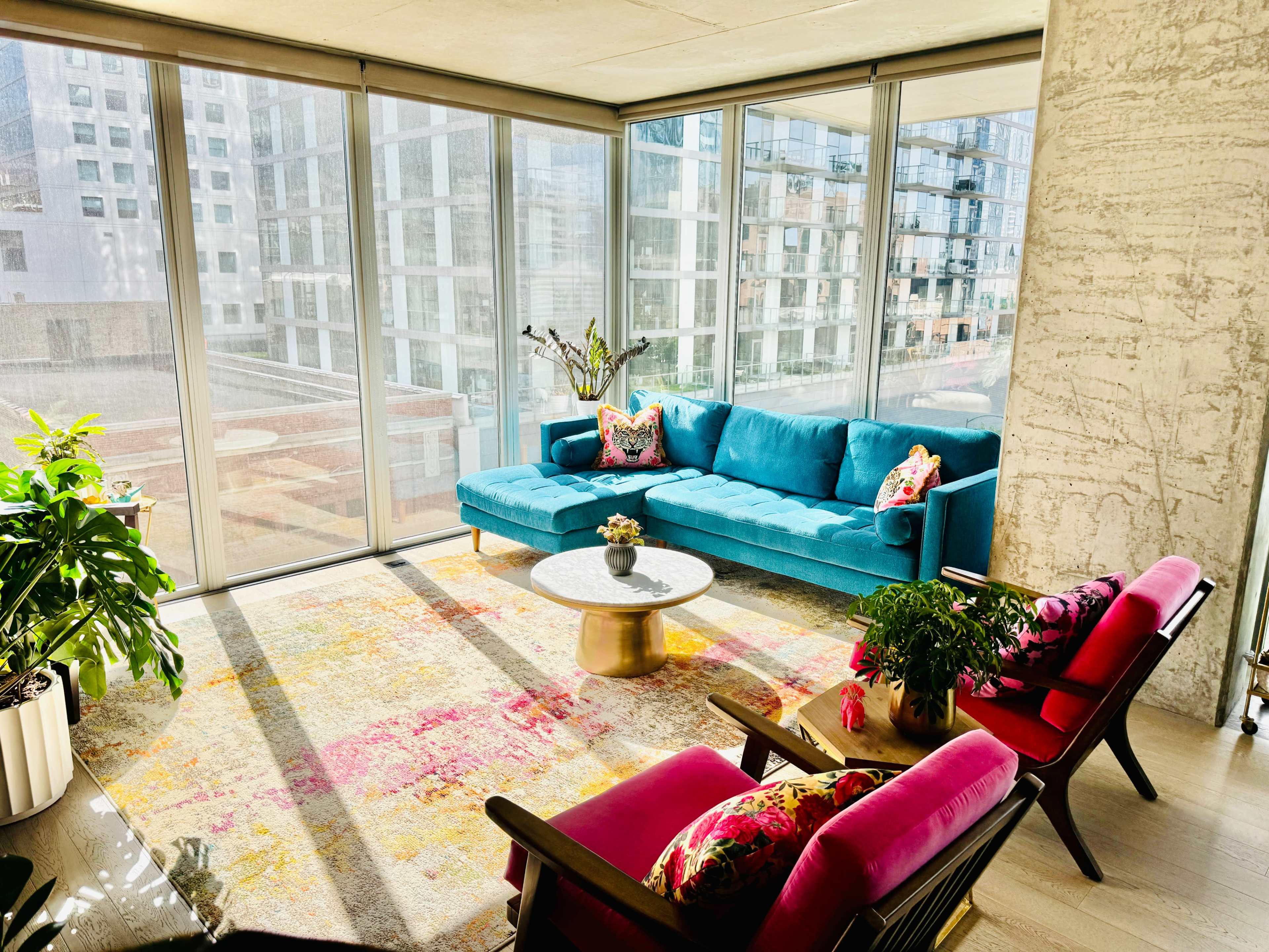 A bright living room features a blue sofa, colorful chairs, a round coffee table, and large windows overlooking a cityscape.