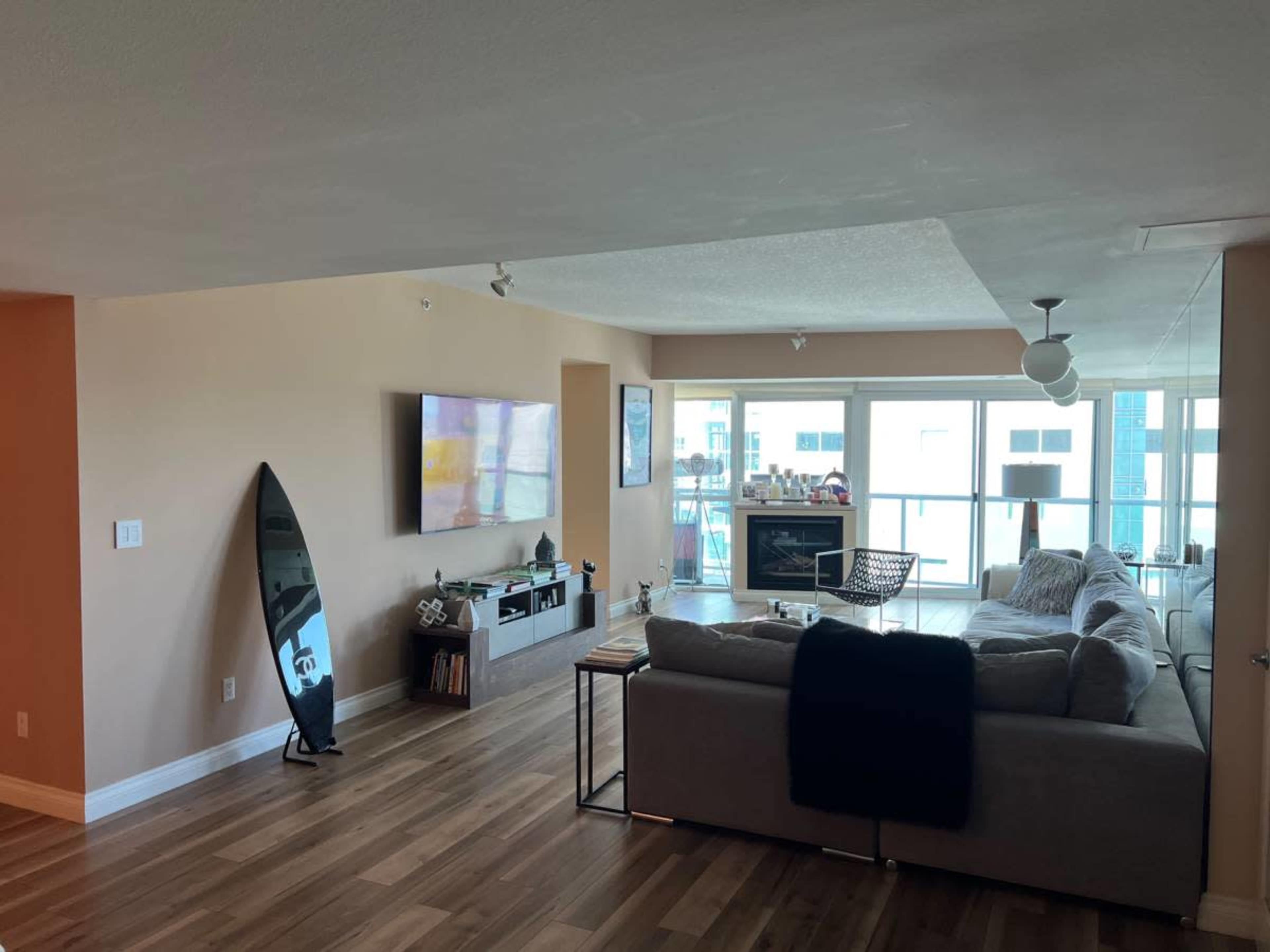 A modern living room features a sectional sofa, a large television mounted on the wall, and a surfboard standing upright in a corner.