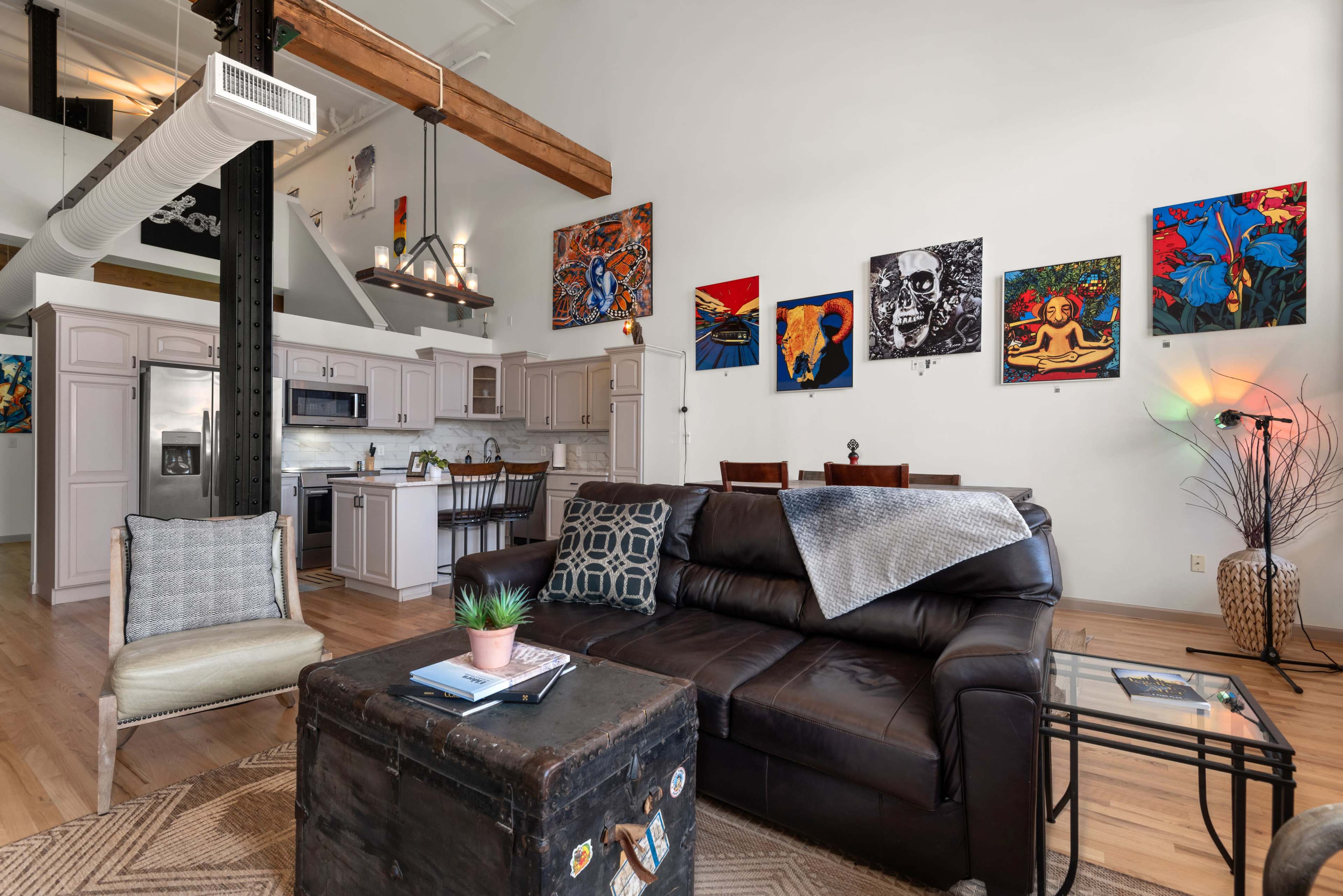 The image shows a spacious living area with a leather sofa, a coffee table, and a kitchen area in the background, adorned with colorful artwork on the walls.