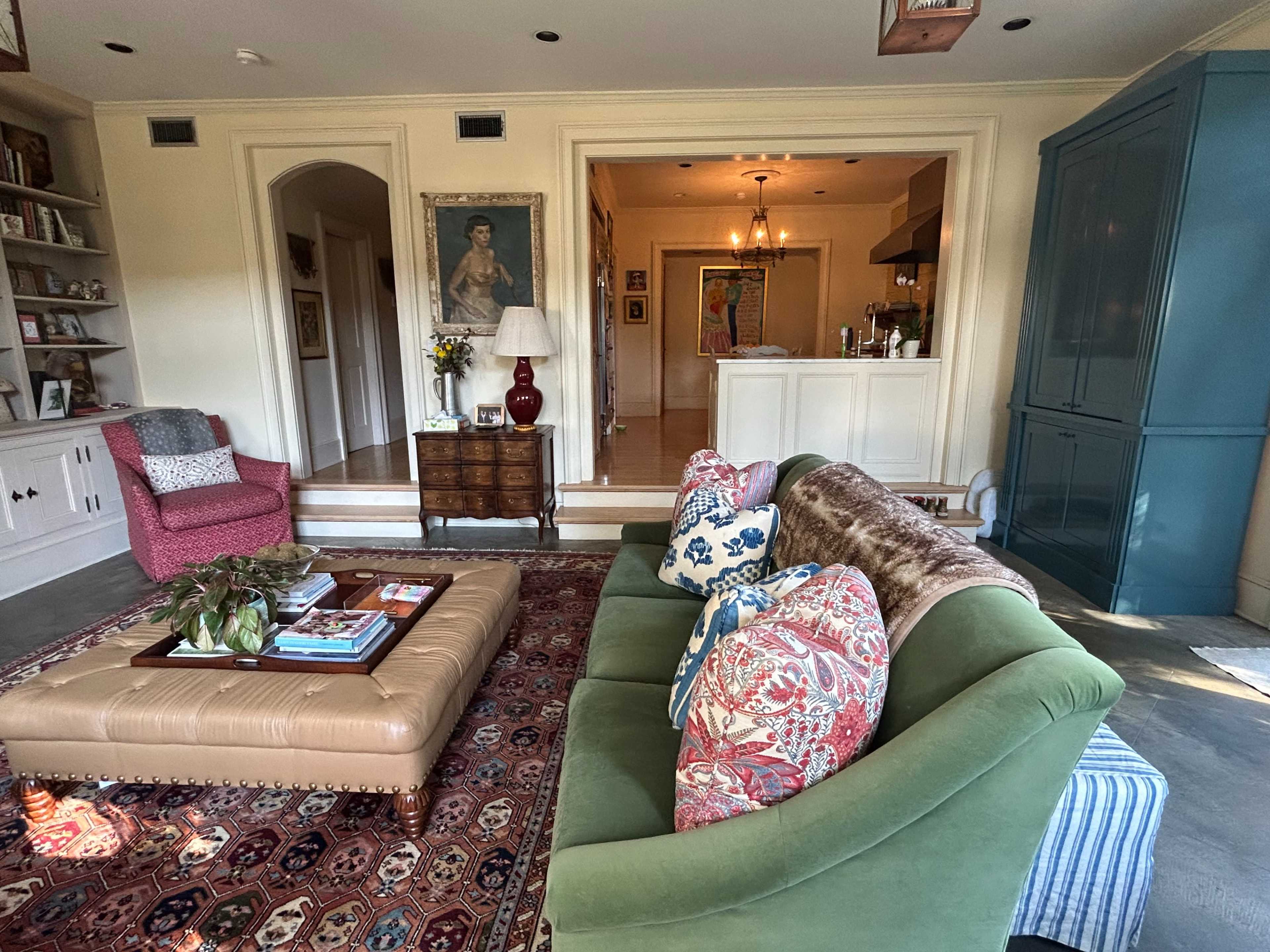The image shows a cozy living room with a green sofa, colorful cushions, a large ottoman, and an adjoining open kitchen area.