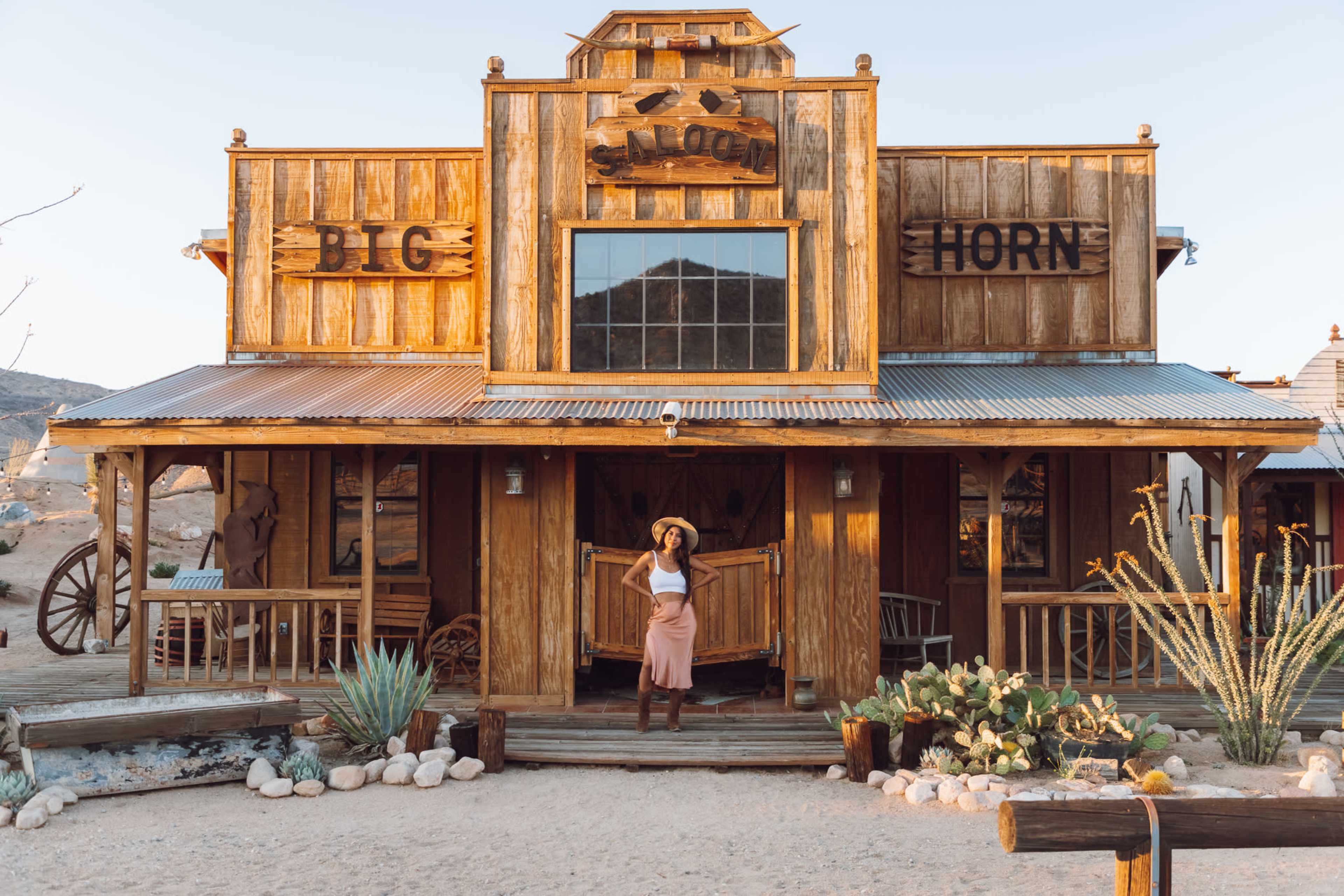 A woman stands at the entrance of a wooden saloon with the sign "BIG HORN" in a desert setting.