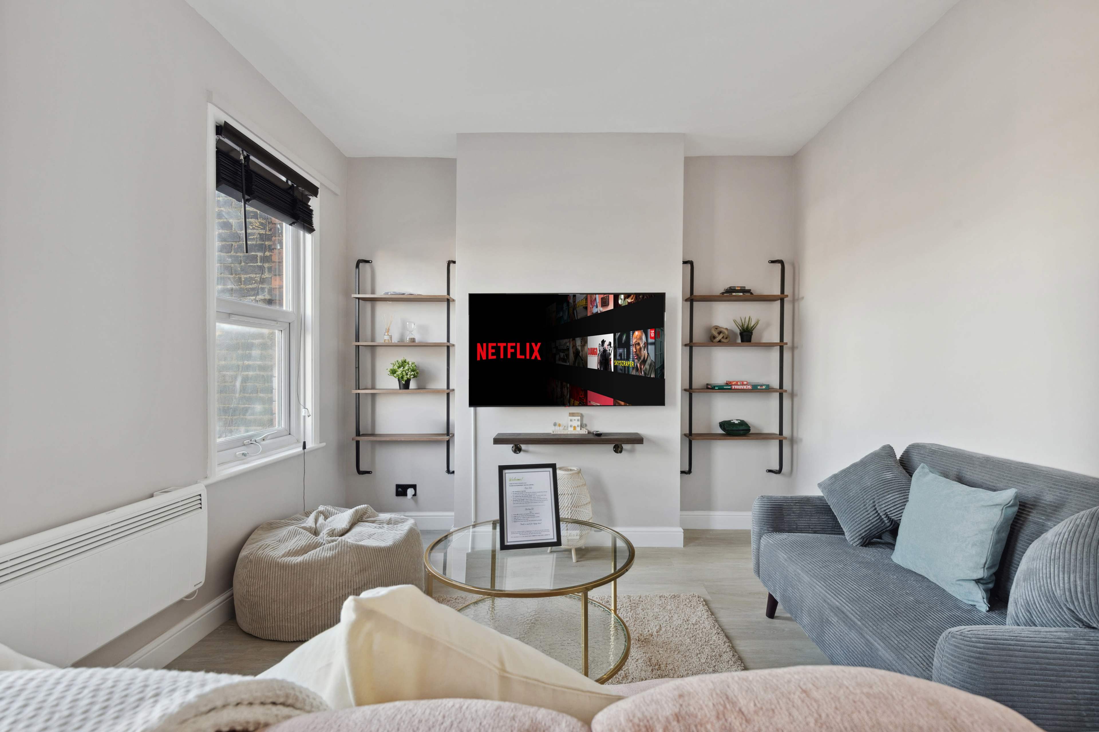 A cozy living room features a wall-mounted TV displaying the Netflix logo, flanked by shelves and furnished with a gray sofa and a round coffee table.