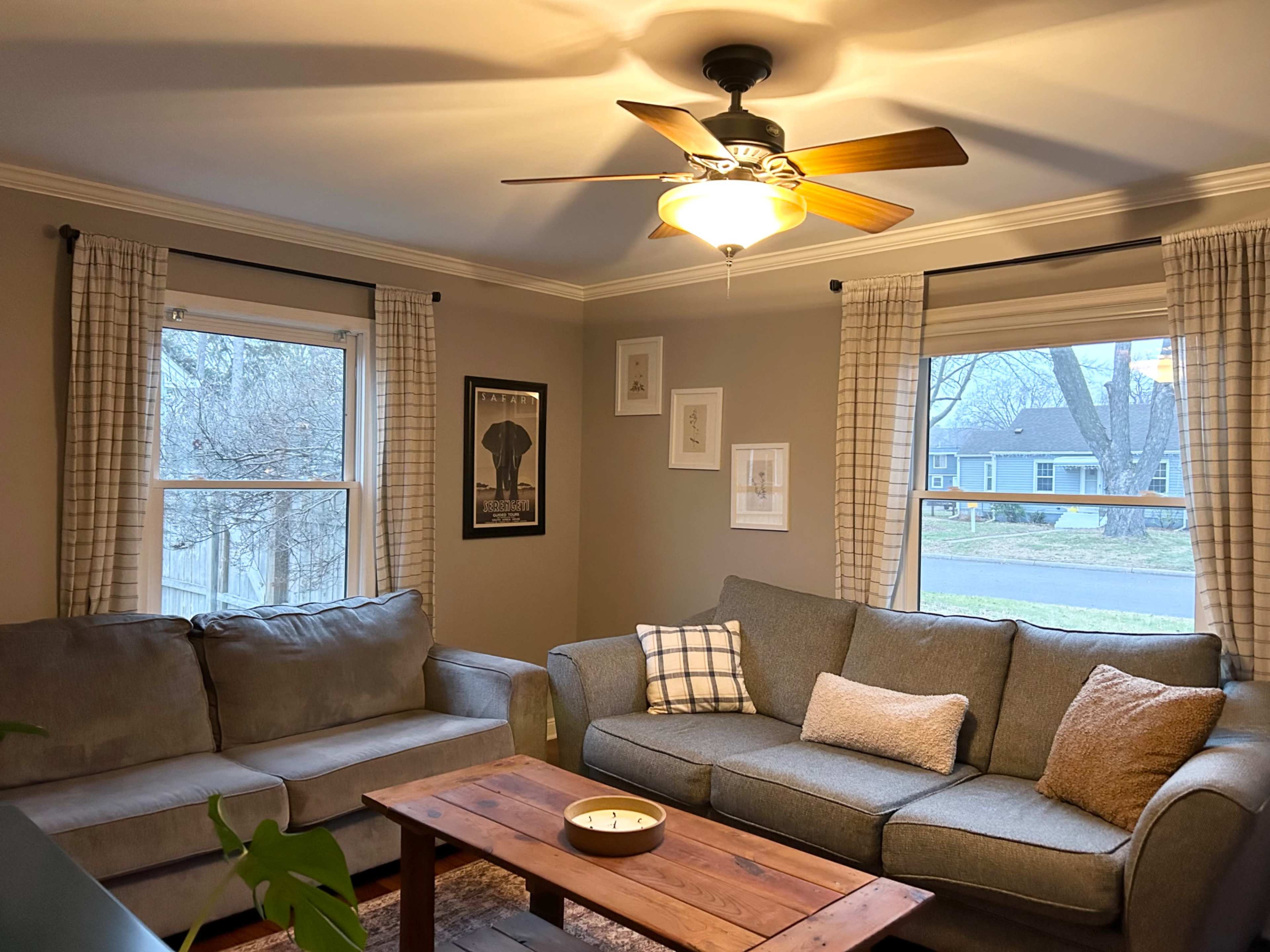 A cozy living room features two gray sofas, a wooden coffee table, a ceiling fan, and framed artwork on the walls beside large windows.