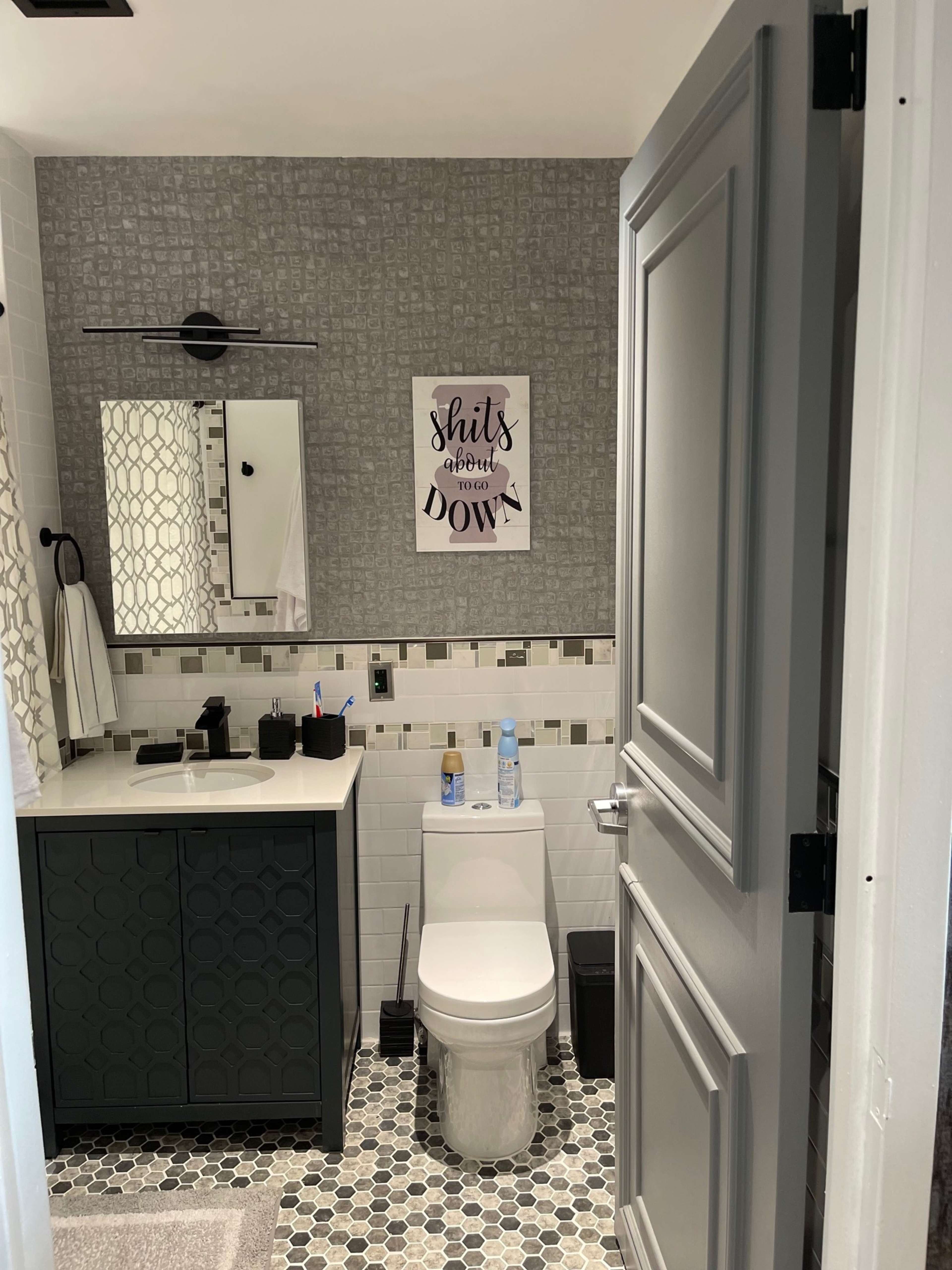 A modern bathroom featuring a toilet, a black vanity with a sink, a mirror, and decorative wall art, all framed by gray walls and patterned tile flooring.