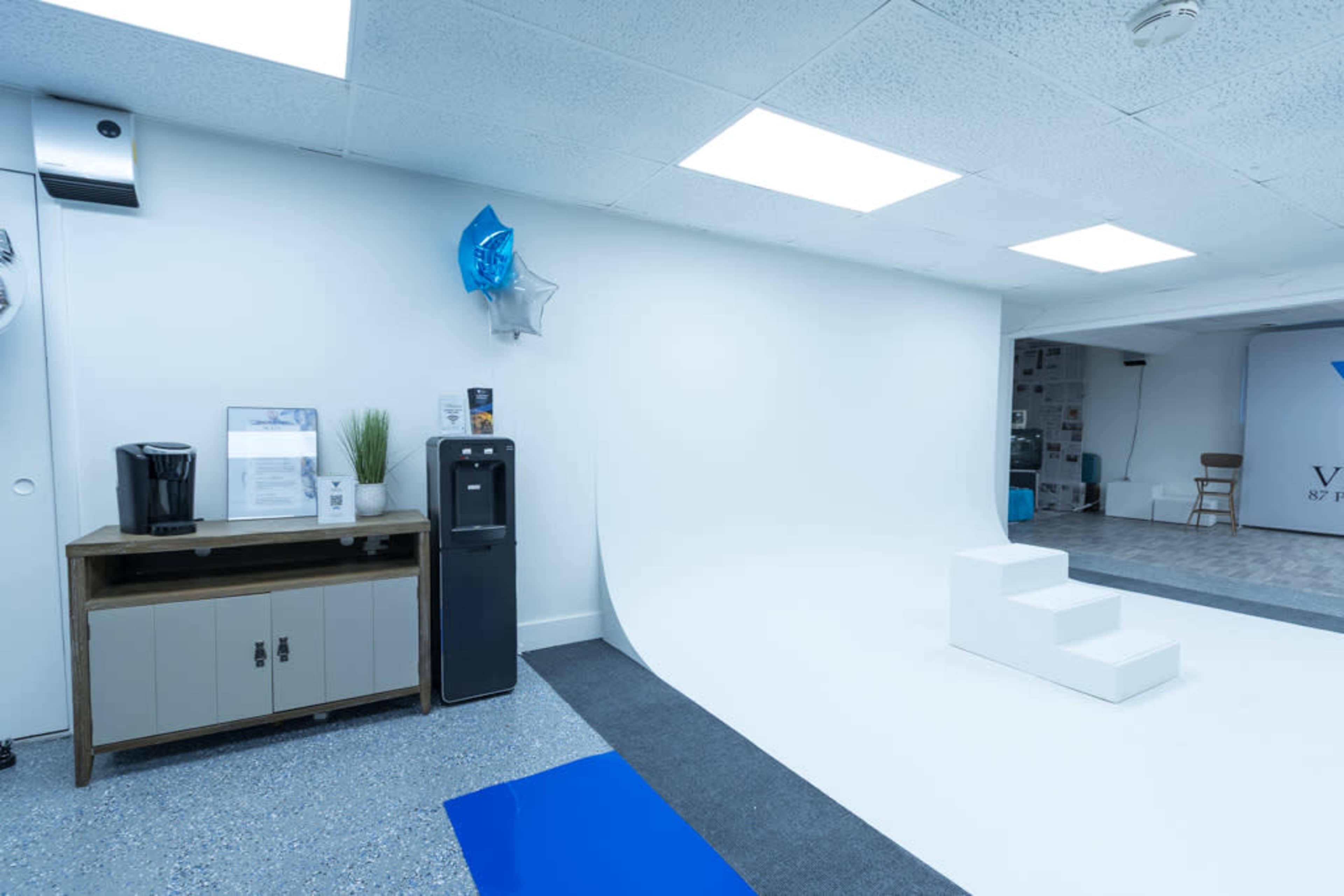 The scene features a minimalistic studio space with a white backdrop, a water cooler, a coffee machine, and a small set of three white steps.