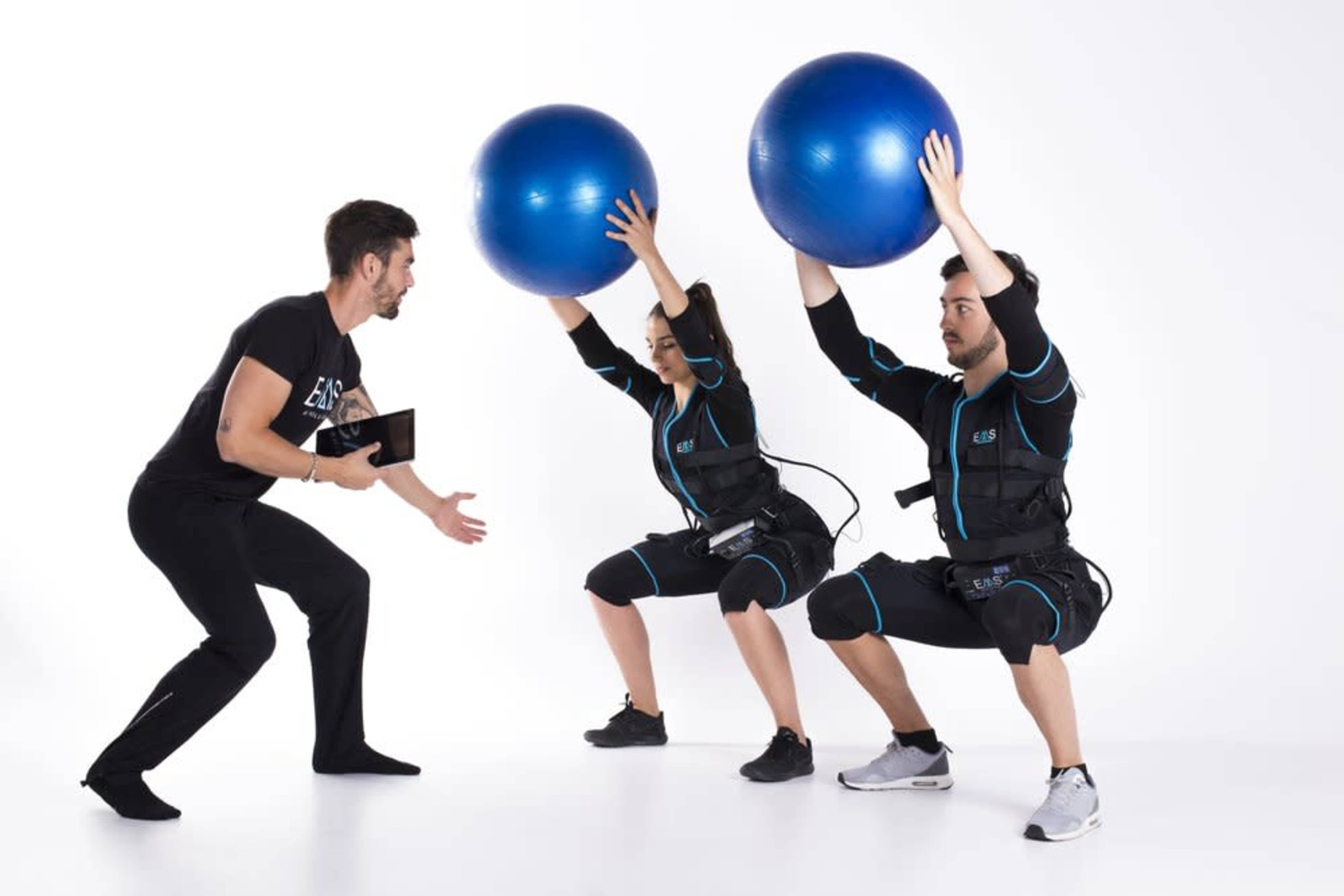 A trainer instructs two individuals in workout attire who are performing squats while holding blue stability balls.