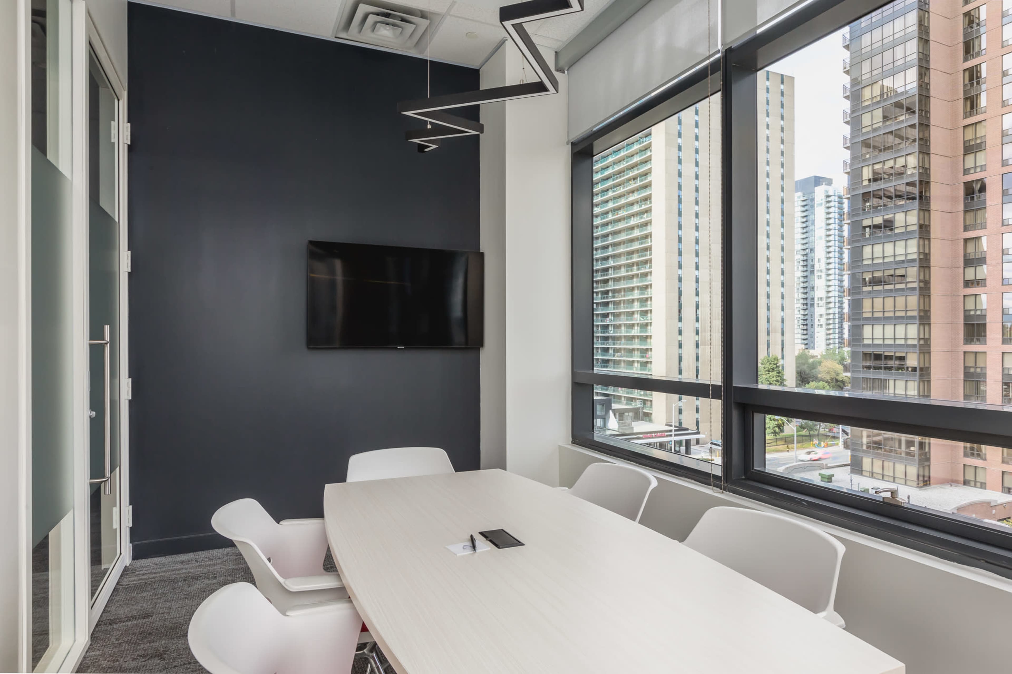 A modern conference room features a long table with white chairs, large windows offering a view of city buildings, and a wall-mounted television.