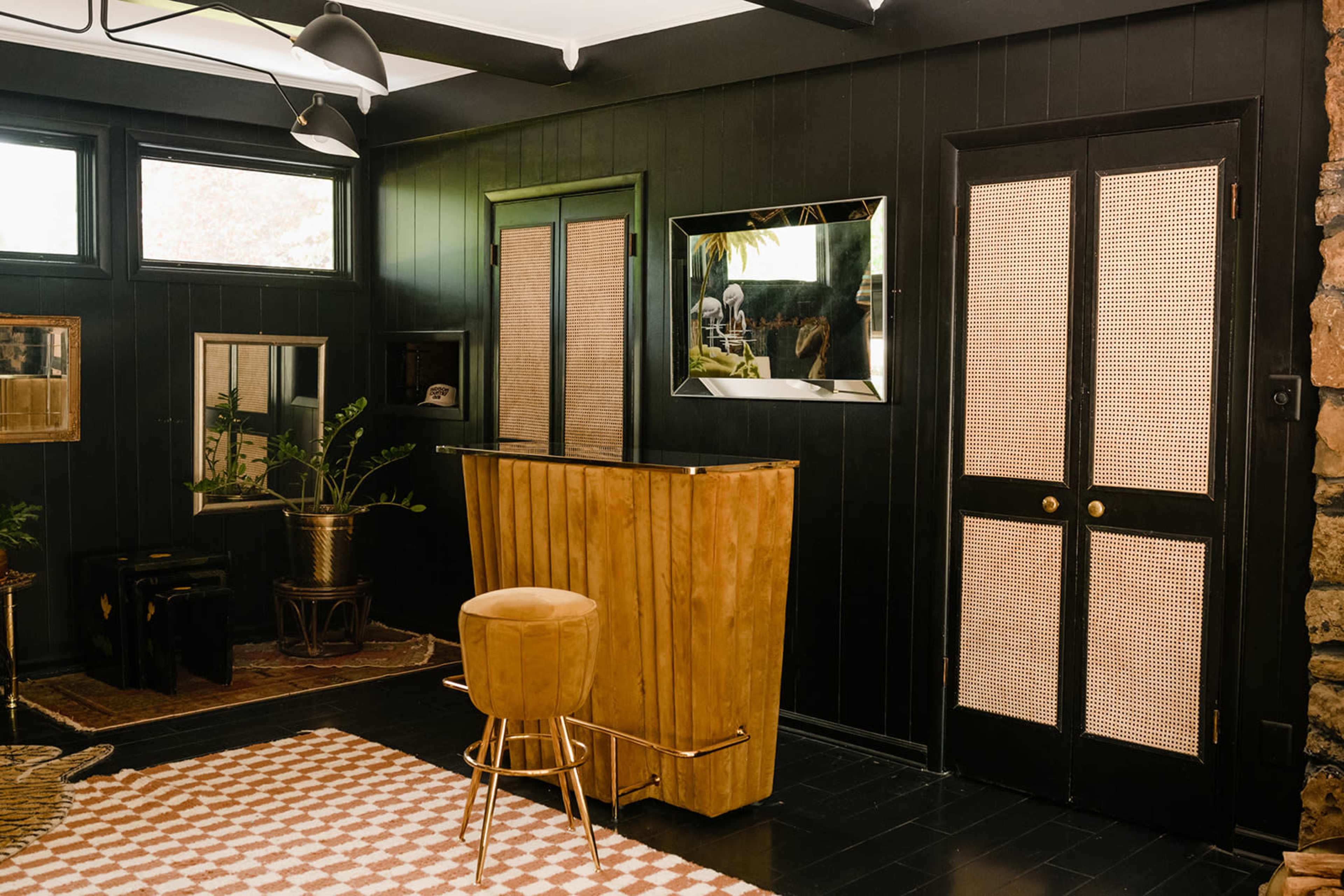 The image shows a dark-colored room featuring a wooden bar with a stool, a large mirror, and doors with woven paneling.