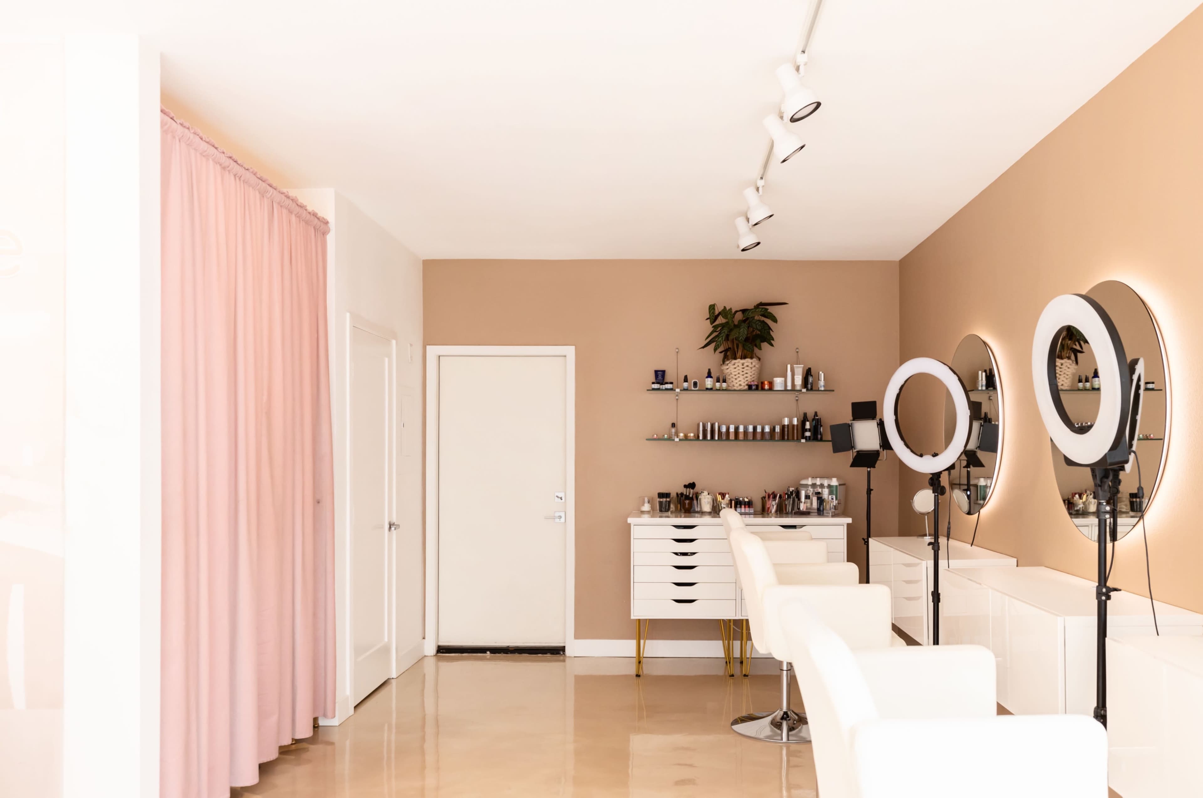 A modern beauty salon featuring white chairs, round mirrors on stands, and a wall decorated with shelves of skincare products.