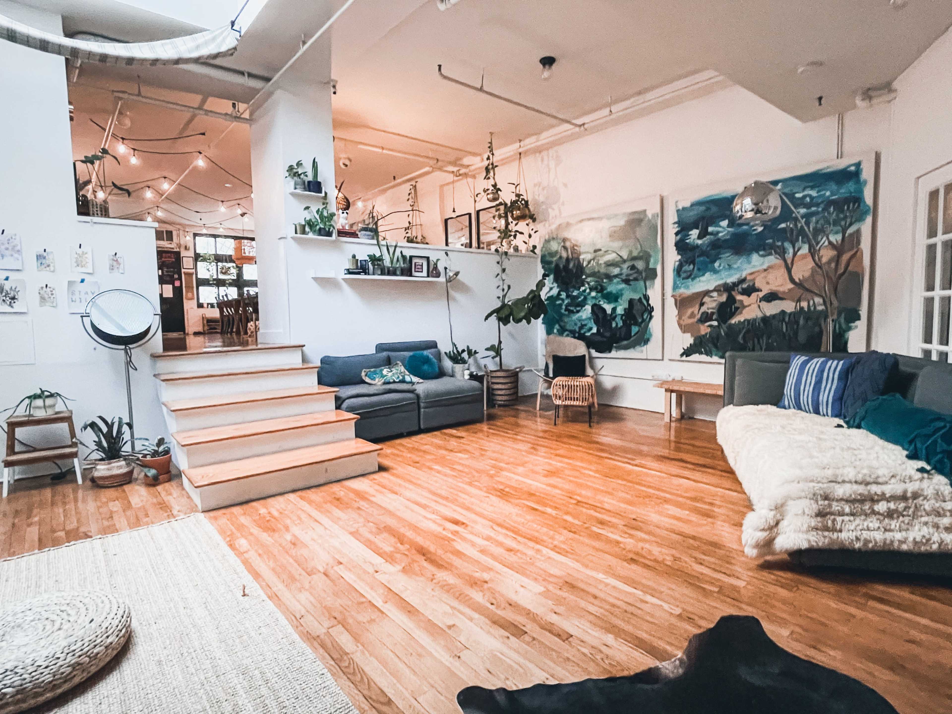 A spacious, well-lit room featuring wooden floors, a stairway leading to a higher level, and several plants arranged throughout the space, with large wall paintings in the background.