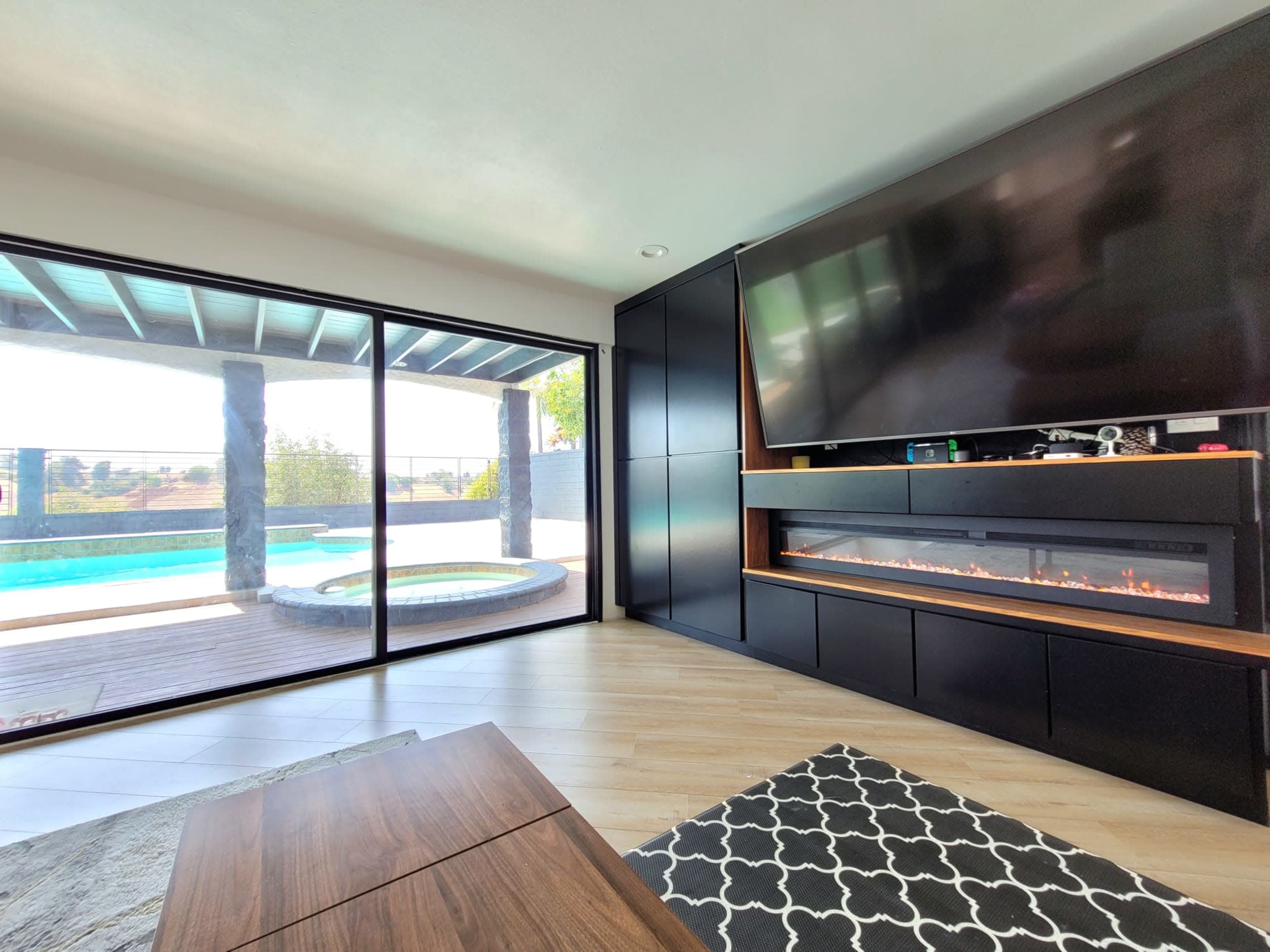 A modern living room with a large television mounted above an electric fireplace, overlooking a pool and outdoor area through expansive glass doors.