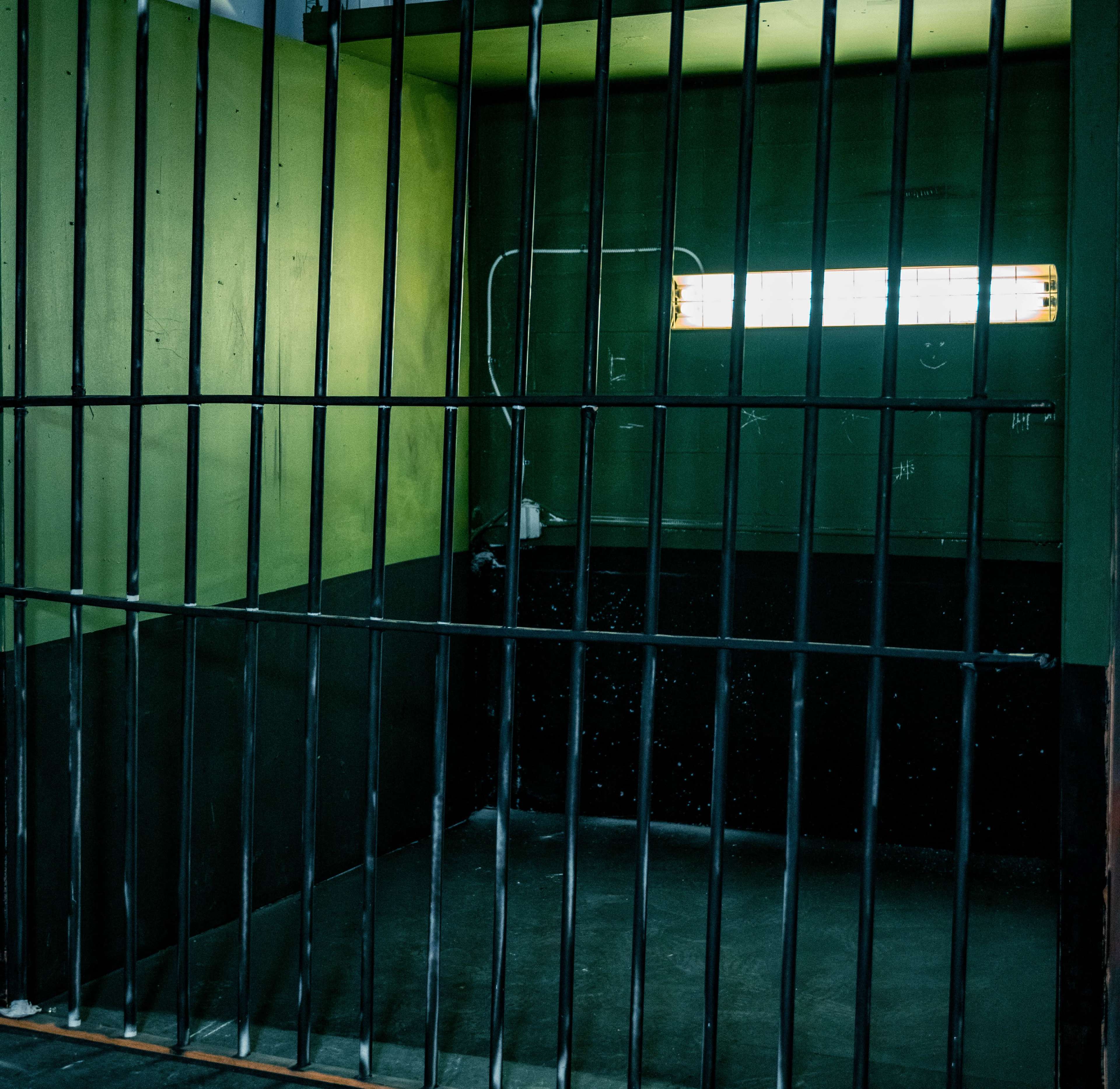 A dimly lit jail cell with black bars and a small window near the top.