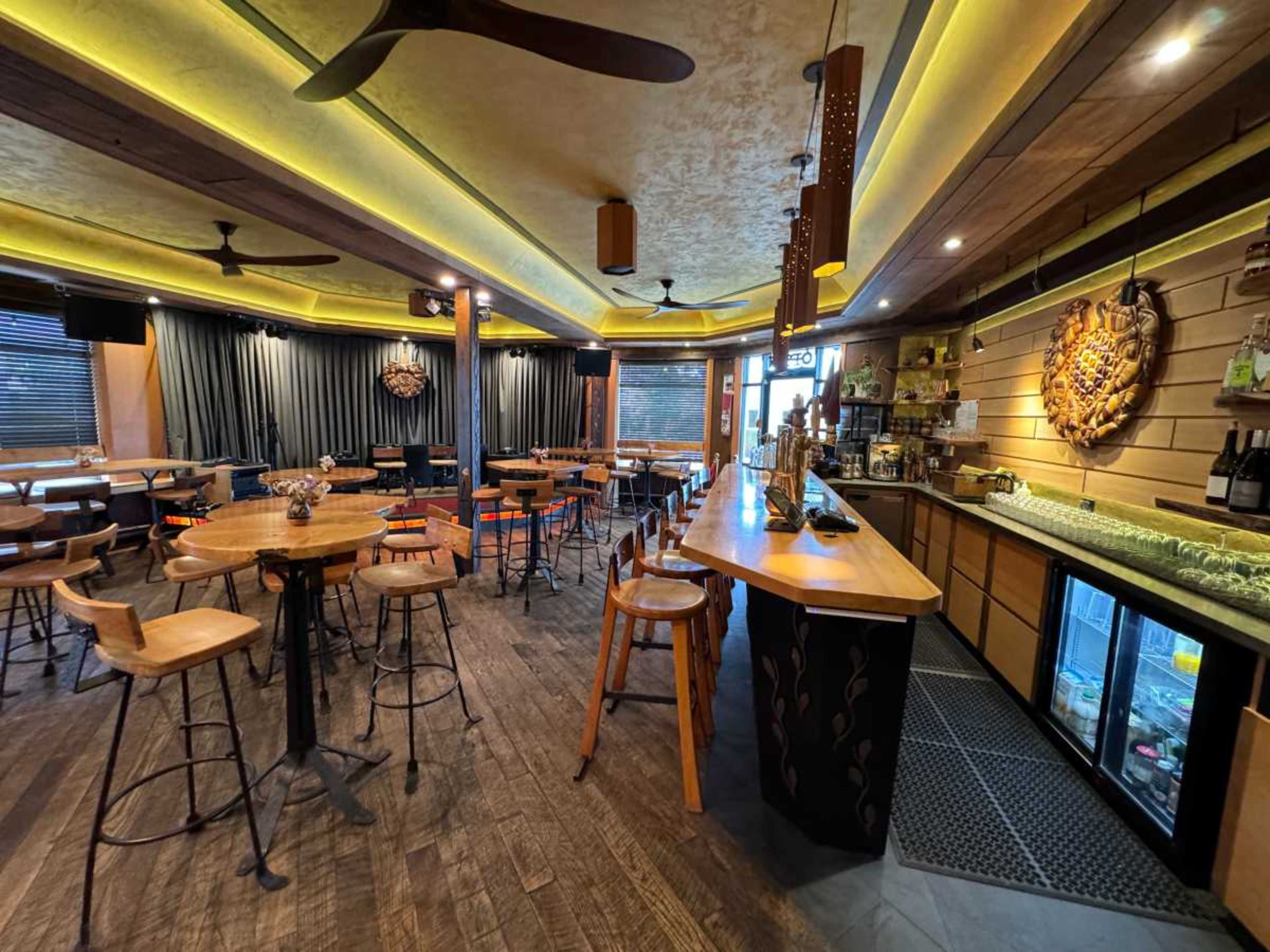 The image shows the interior of a restaurant or bar featuring wooden tables, stools, and a well-lit counter with a drinks fridge.