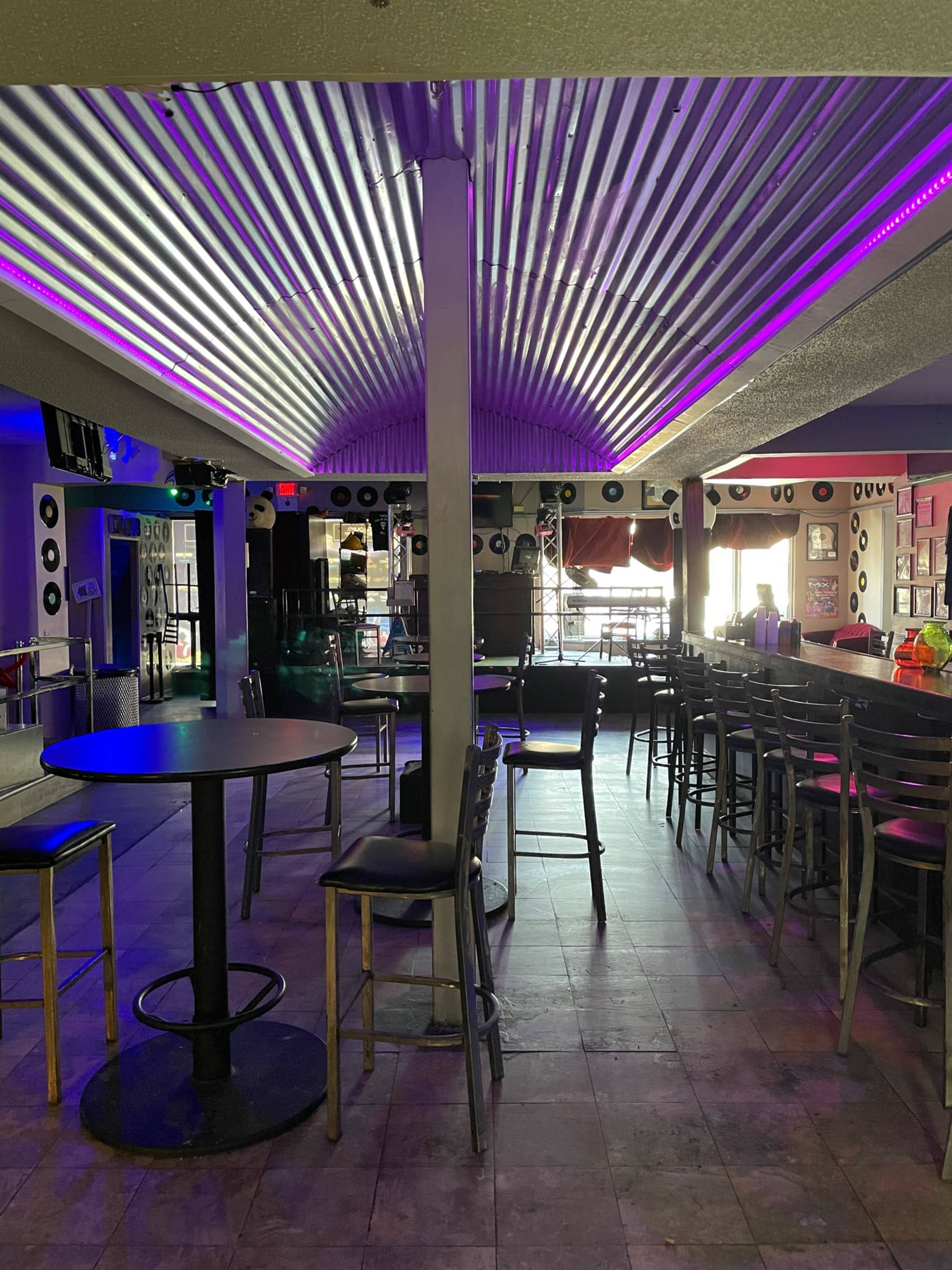 A half-empty bar with metal siding on the ceiling, vibrant purple lighting, and empty tables and chairs.