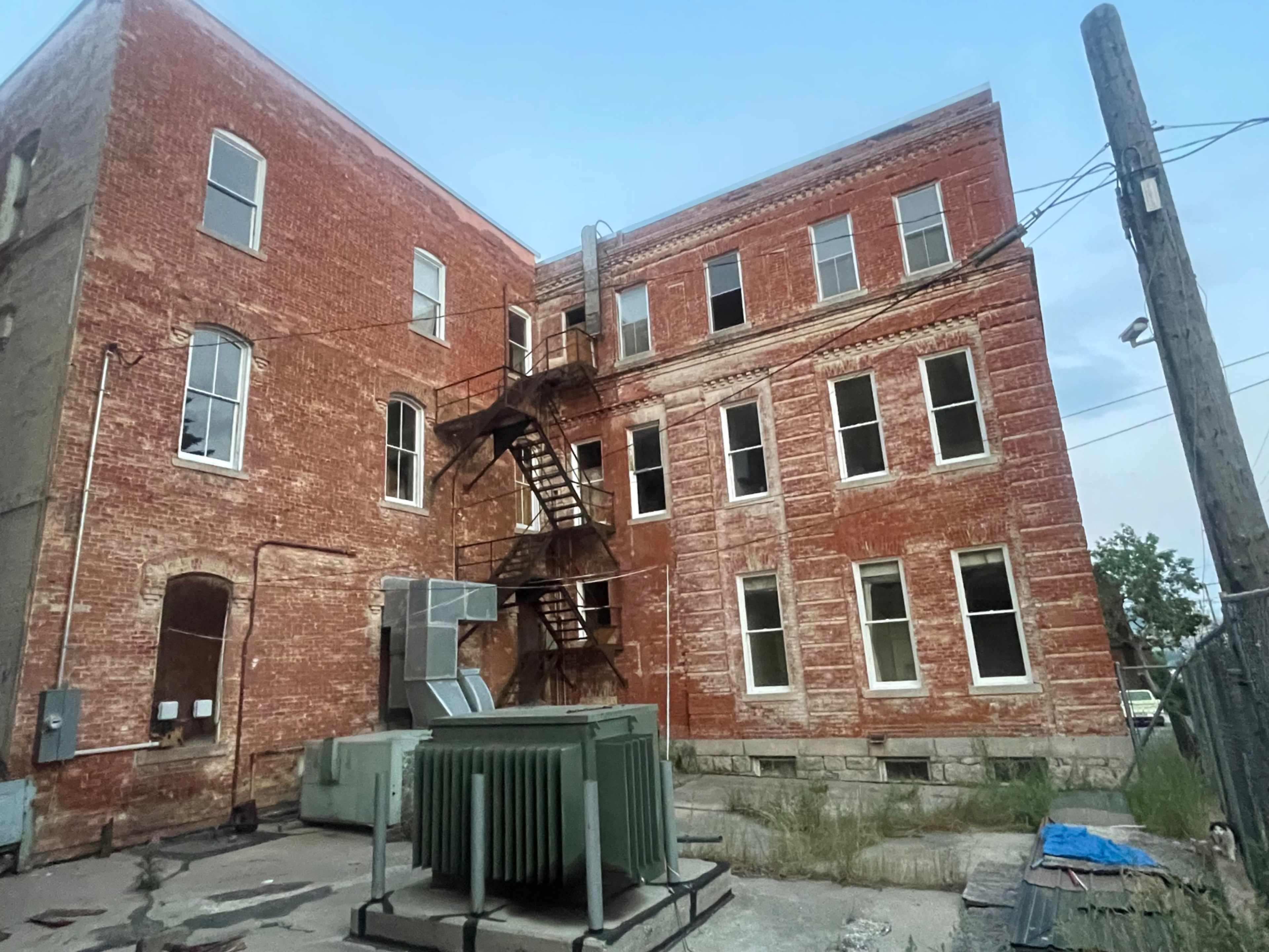 The image shows a weathered brick building with a metal fire escape, abandoned air conditioning units, and broken windows.