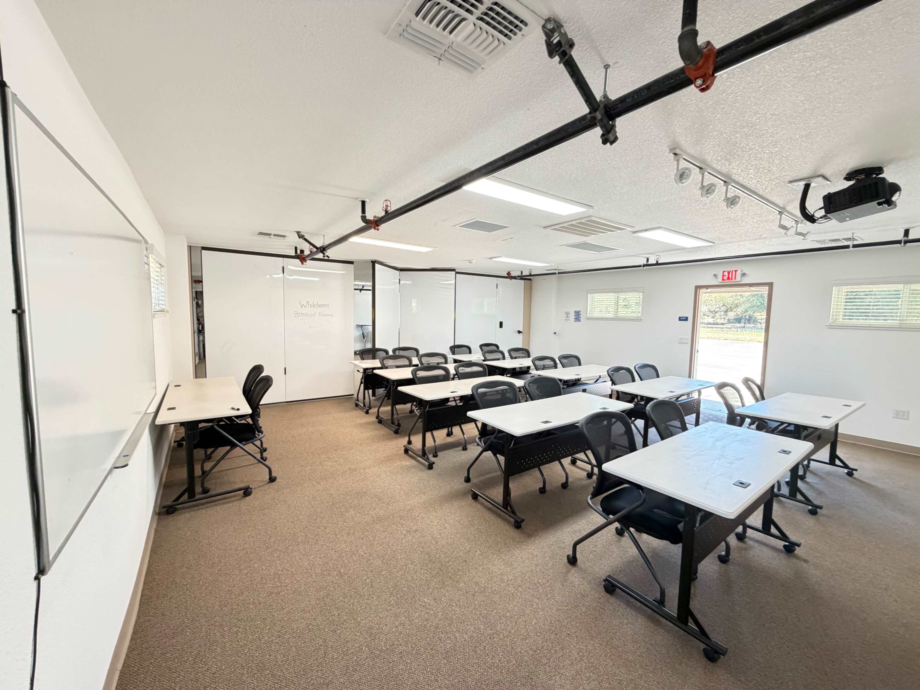 A classroom is set up with rows of desks and chairs, a whiteboard, and a projector, all under a bright overhead light.