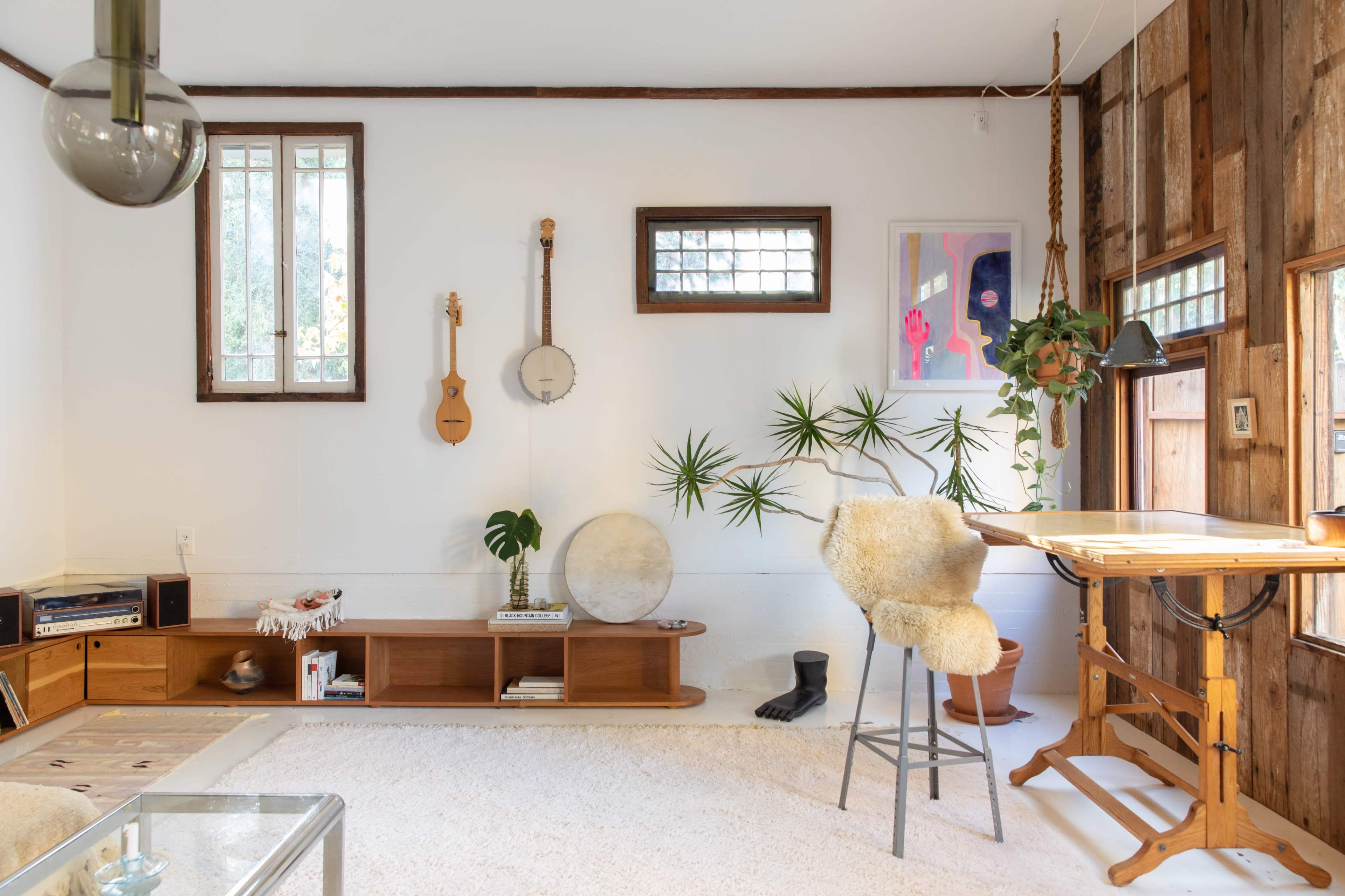 A cozy interior features a wooden shelf with various décor items, a chair at a table, plants, and art on the walls.