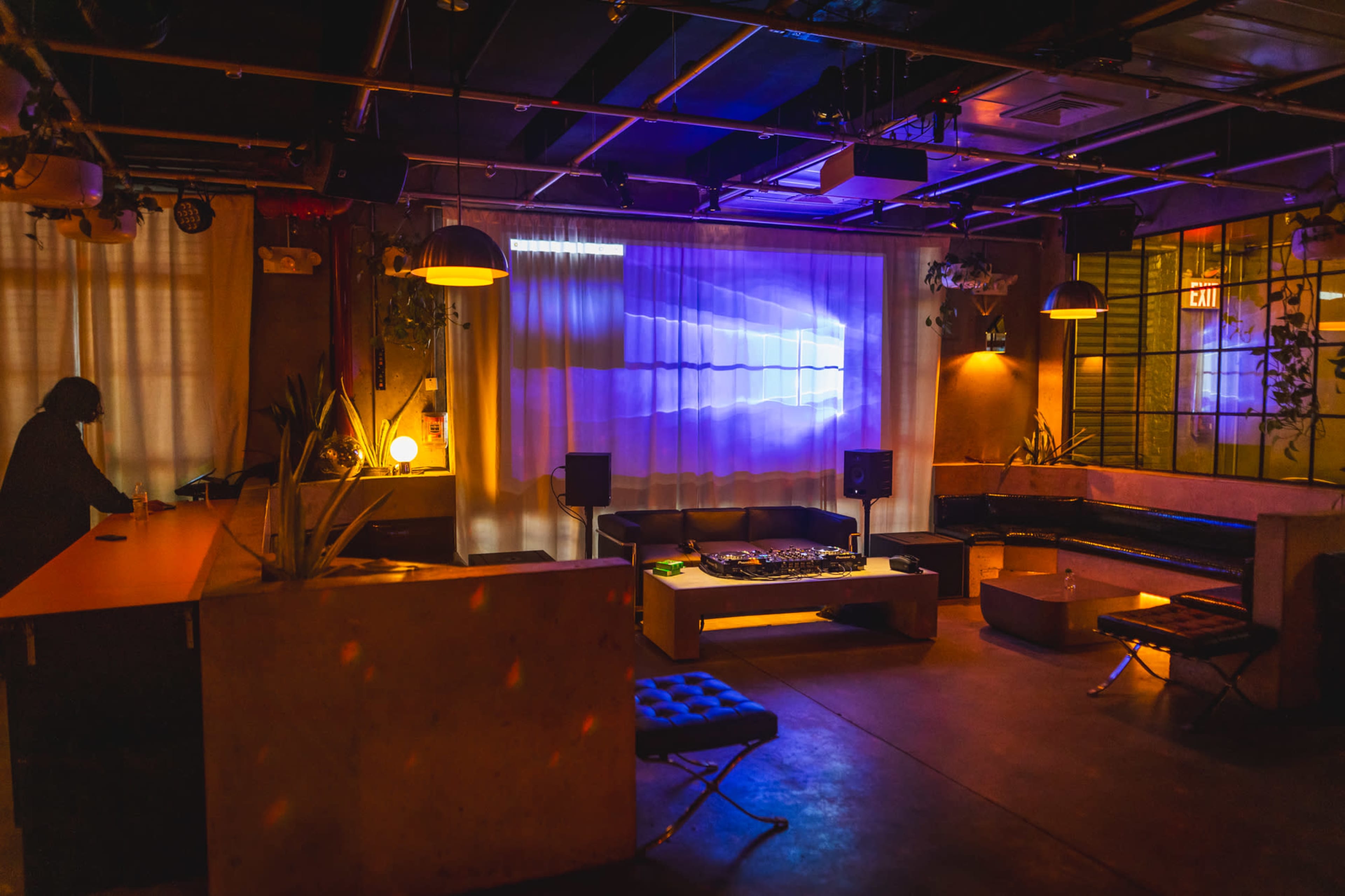 A dimly lit lounge area features a DJ booth, seating, and ambient lighting with a projection on the wall.