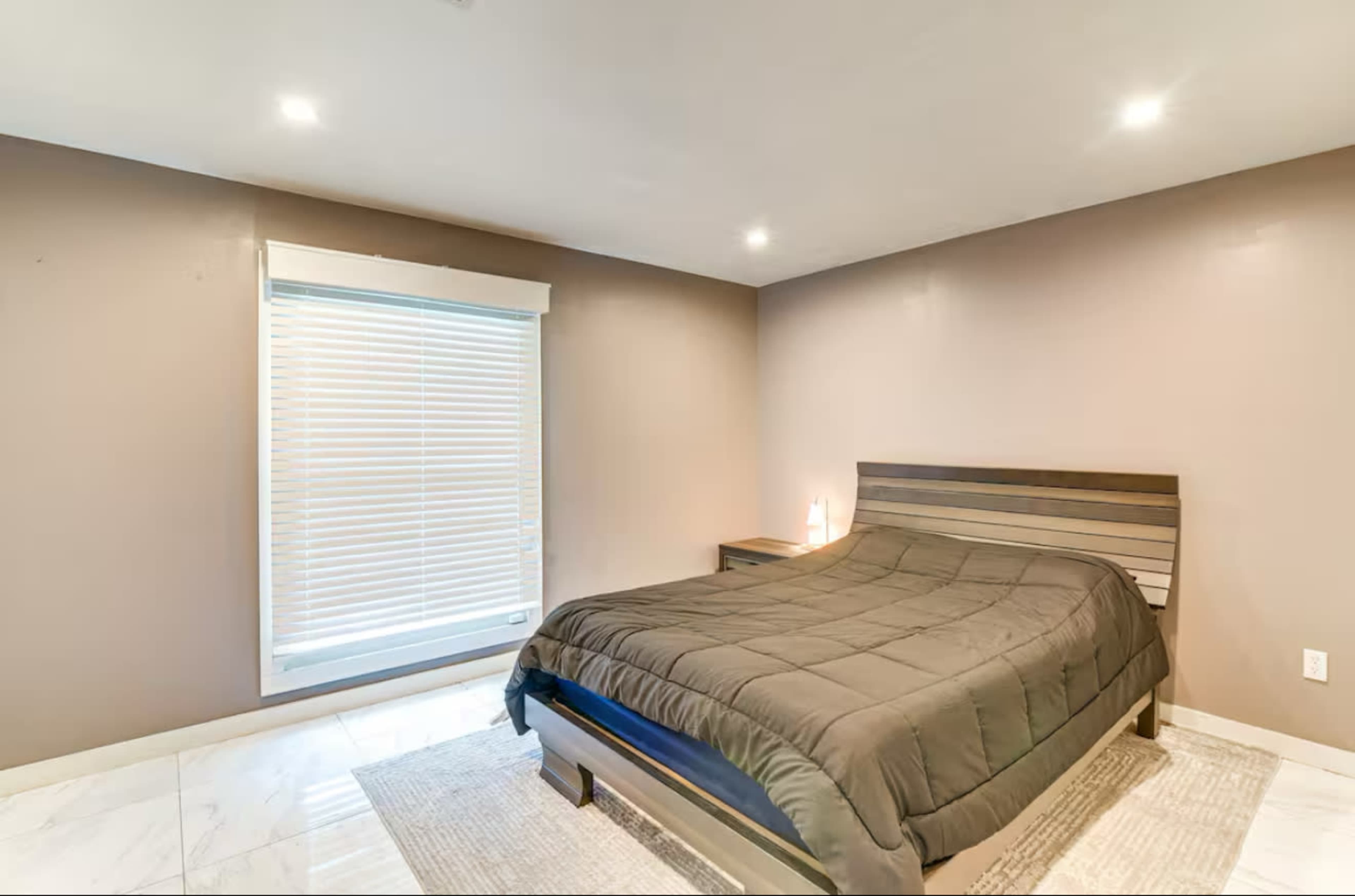 A minimalistic bedroom features a large bed with a dark comforter, a nightstand, and a window with closed blinds.