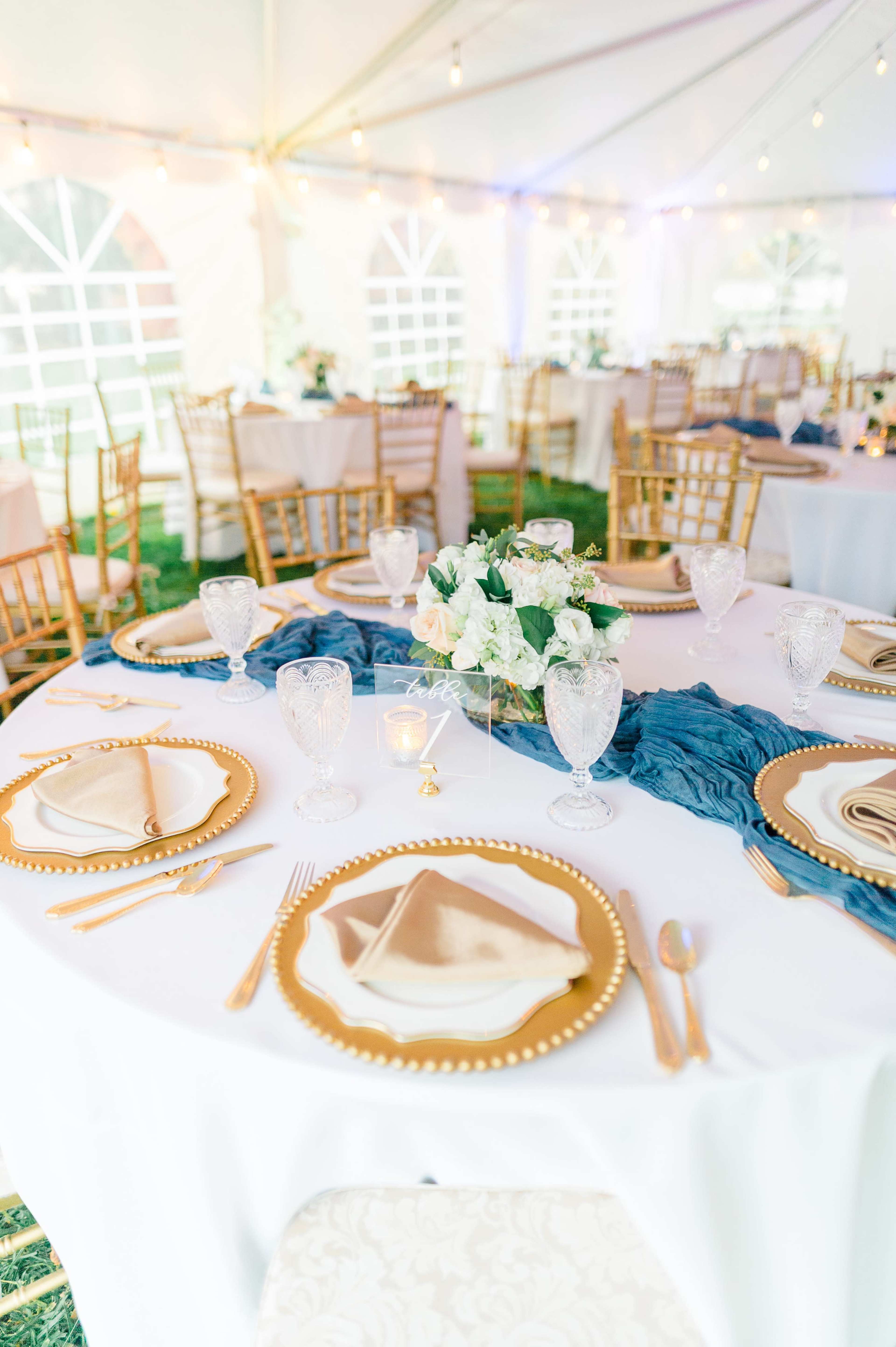 The image shows a beautifully arranged dining setup under a tent, featuring round tables with white tablecloths, gold-rimmed plates, crystal glassware, and floral centerpieces.