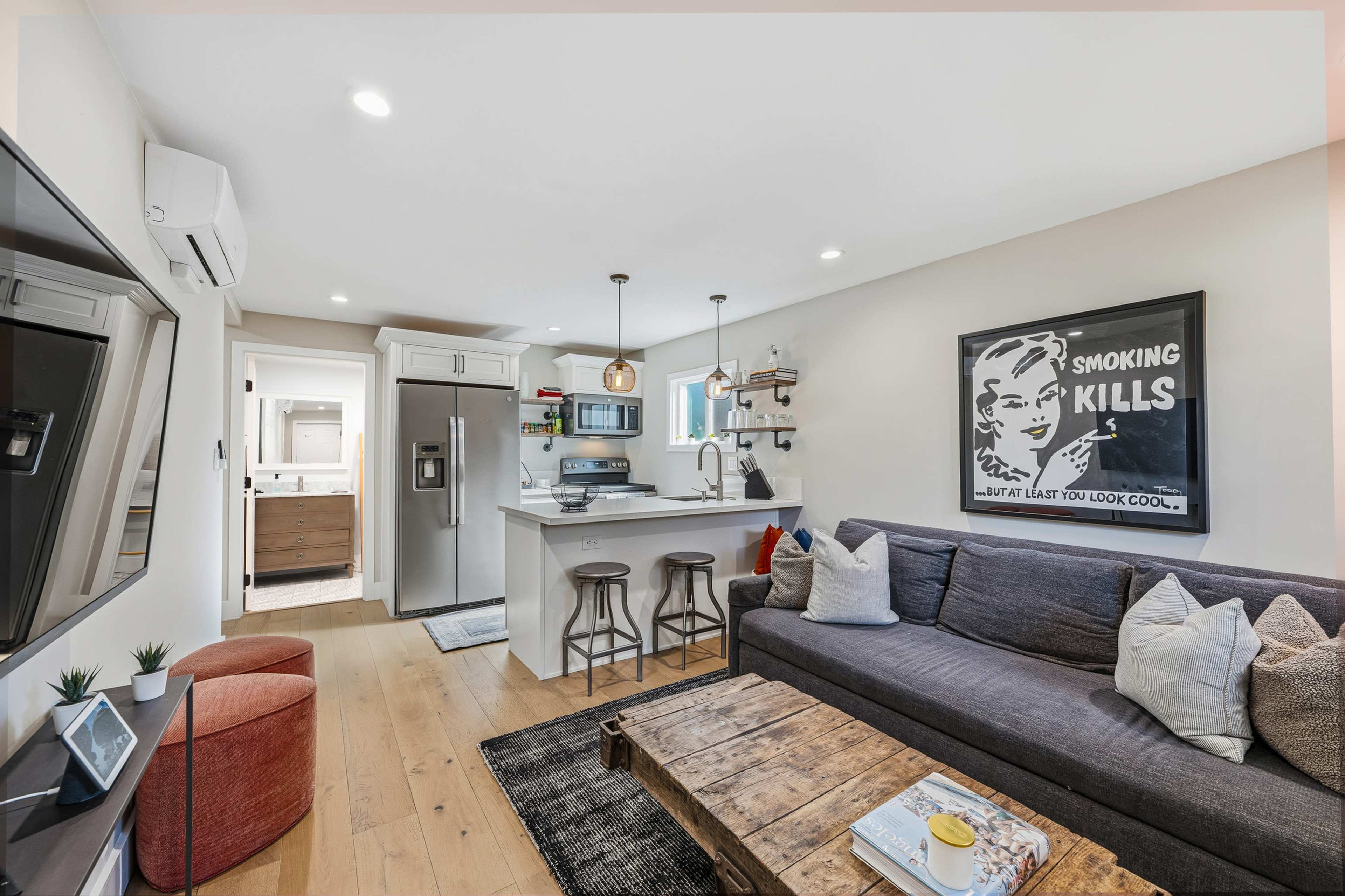 The image shows a modern, open-concept living space featuring a gray sofa, a wooden coffee table, and a kitchenette with stainless steel appliances.