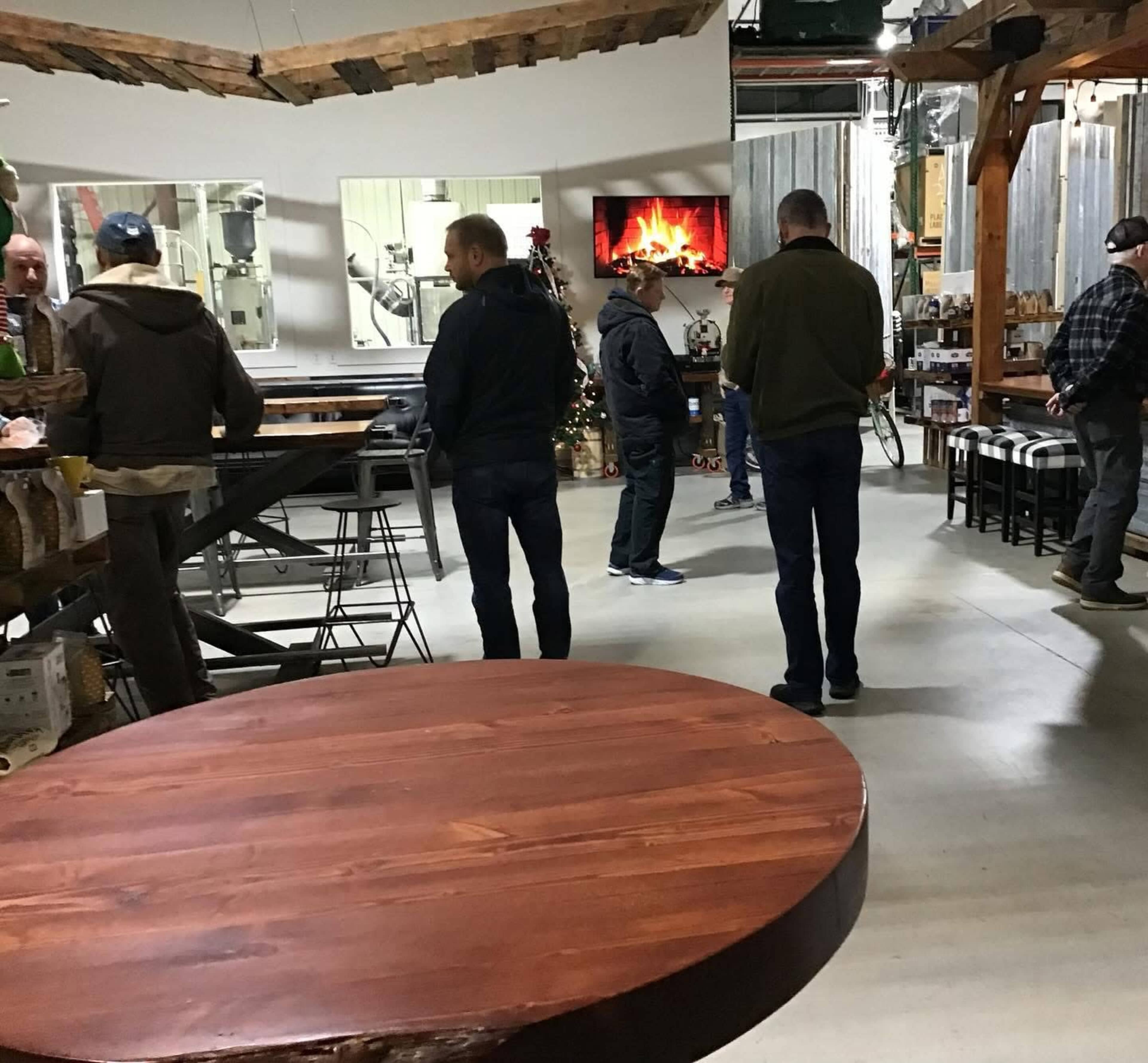 A group of people stands in a spacious industrial-style room with a large wooden table and a digital fireplace display on the wall.