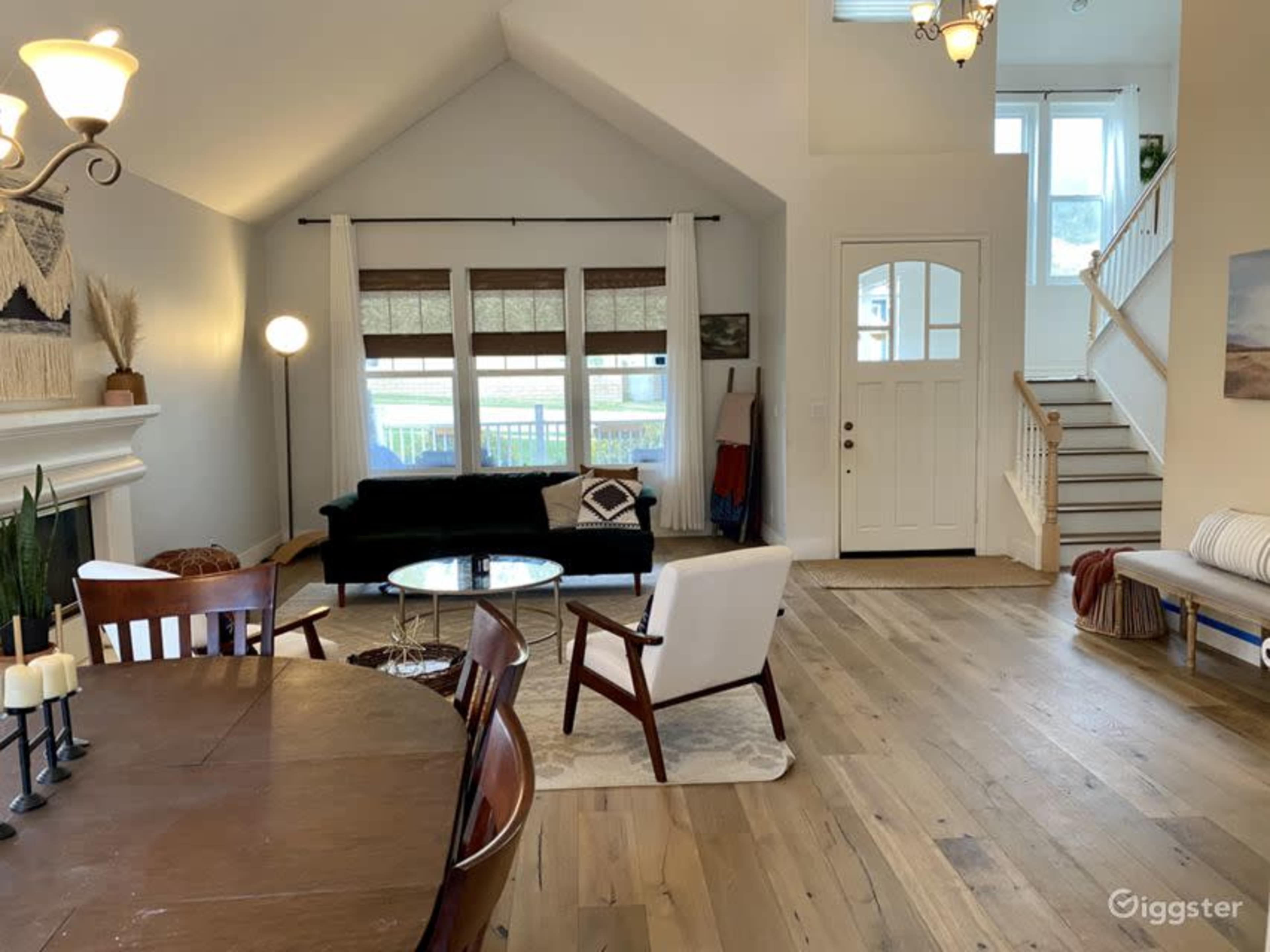 The image shows a bright, spacious living room with a mixed seating arrangement, large windows, and an entryway leading to a staircase.