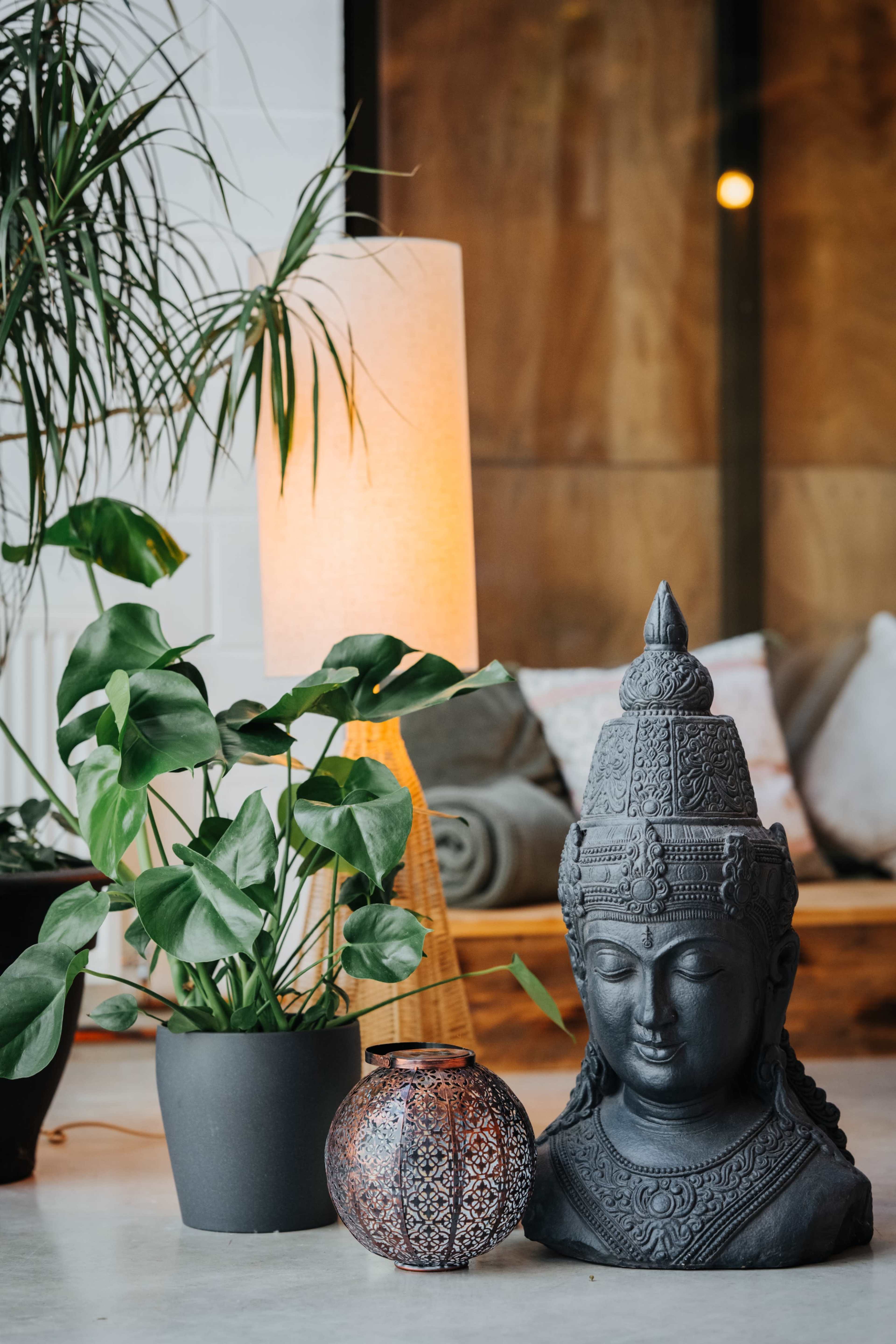 A black Buddha statue sits beside a potted plant and an ornate lantern, with a floor lamp illuminating the scene in a cozy interior space.