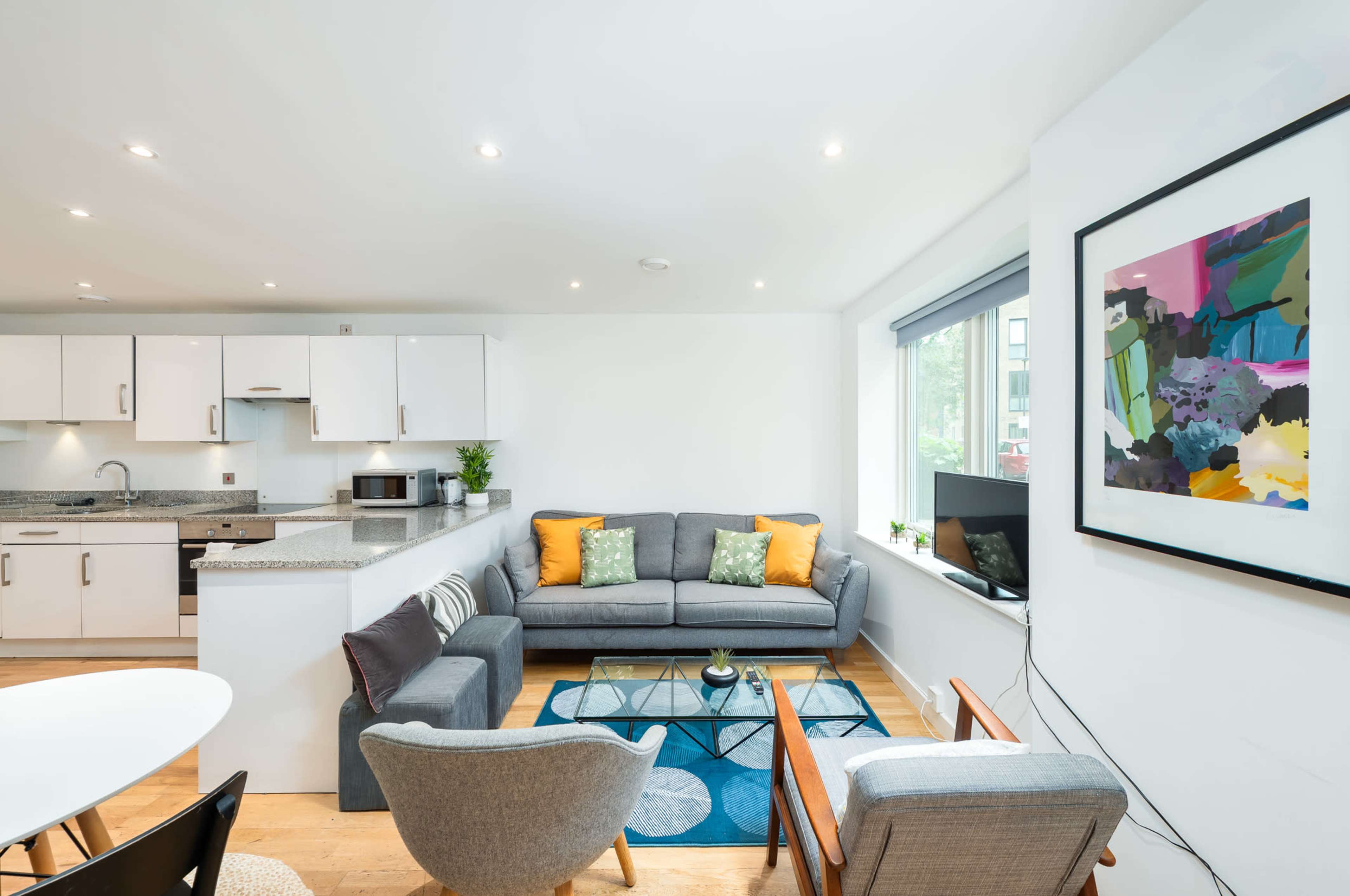 The image shows a modern, open-plan living area with a gray sofa, patterned rug, a coffee table, and a kitchen with white cabinetry in the background.