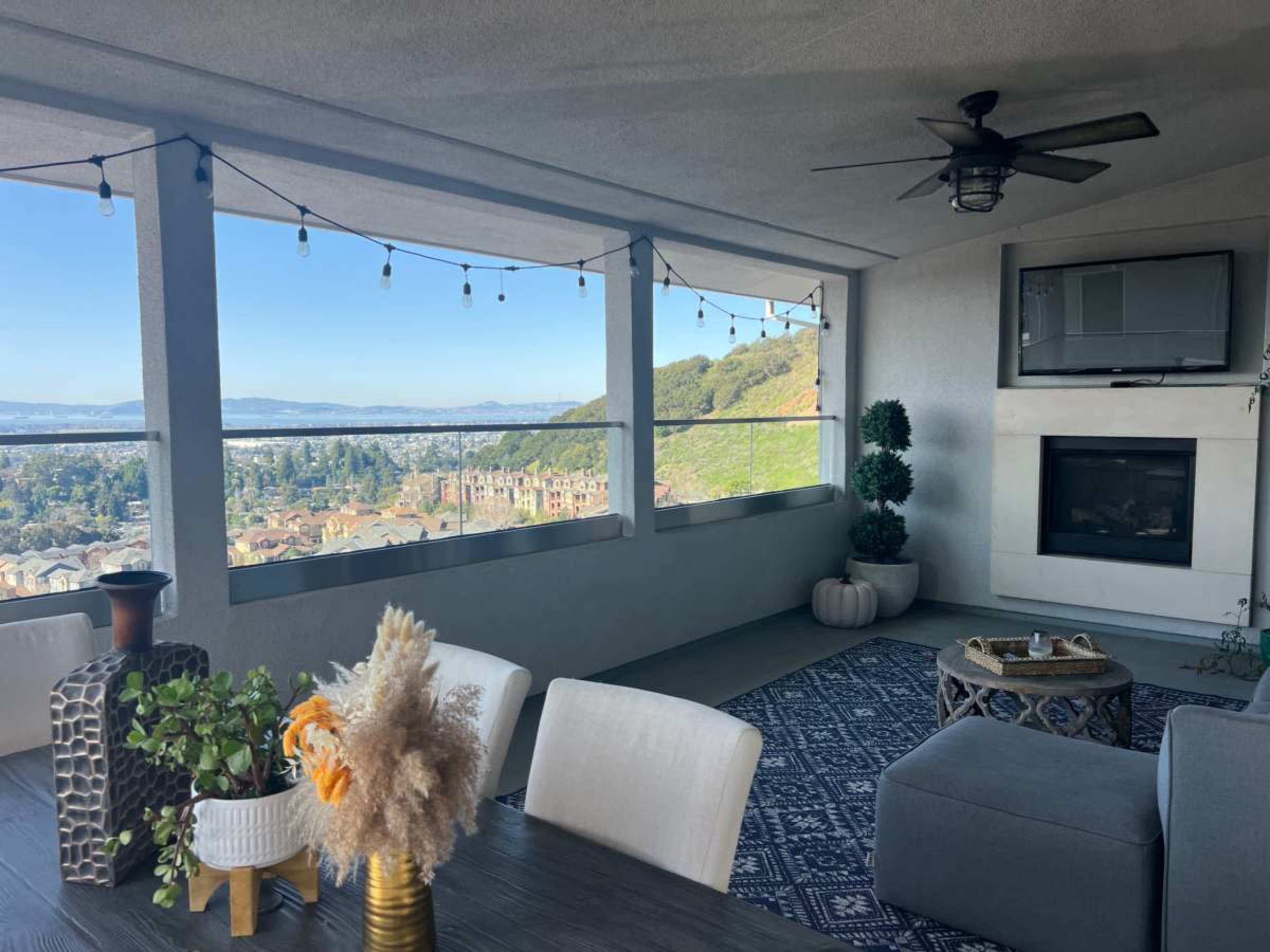 A modern outdoor patio features a dining table, decorative plants, and a view of rolling hills and a distant cityscape.