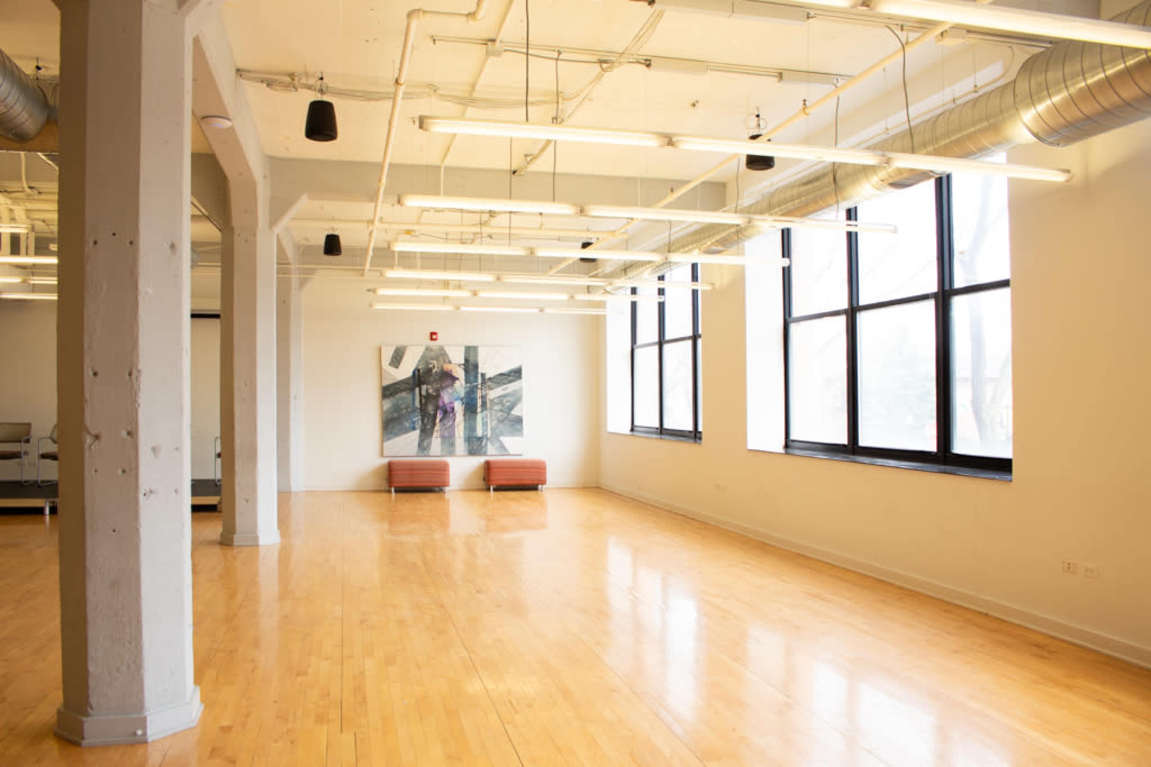 The image shows a spacious, well-lit interior with wooden flooring, large windows, and minimal furnishings, including two orange couches and a wall-mounted artwork.