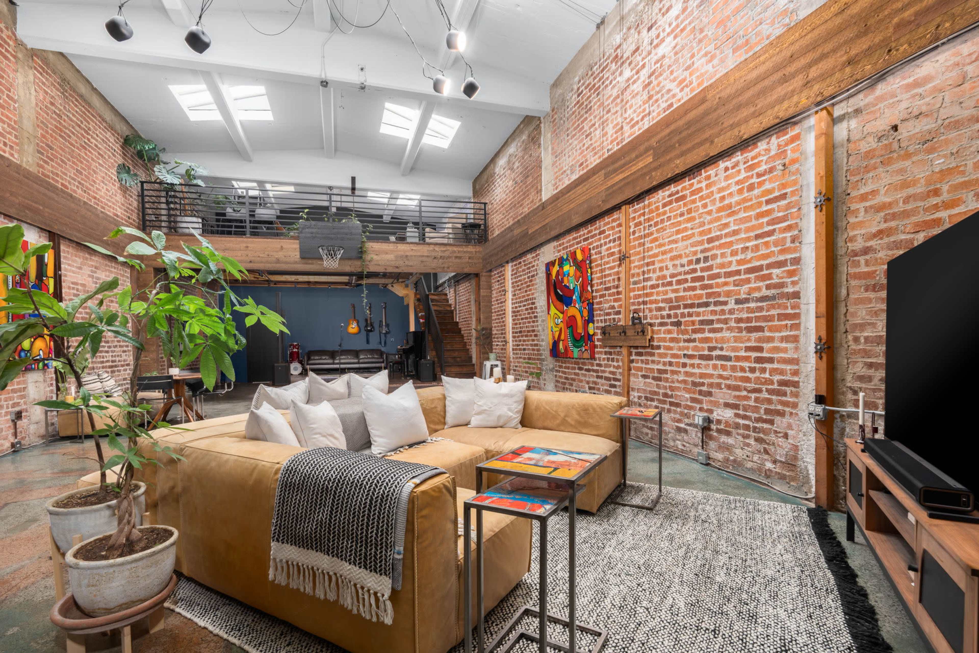 A spacious loft with exposed brick walls, a large yellow sectional sofa, and a basketball hoop visible in the background.