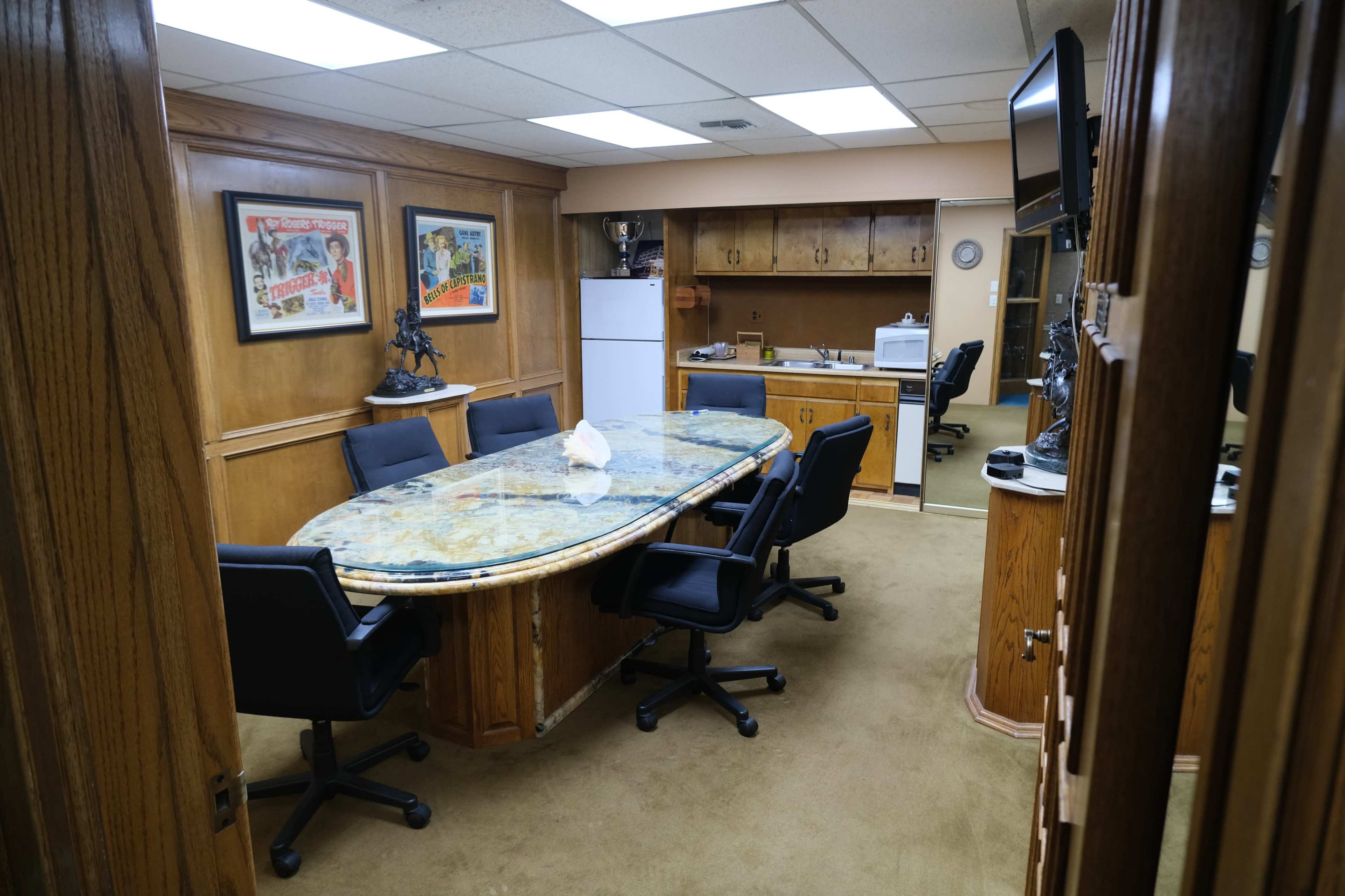 A conference room with a large oval table surrounded by black chairs, featuring wood-paneled walls and a refrigerator in the background.