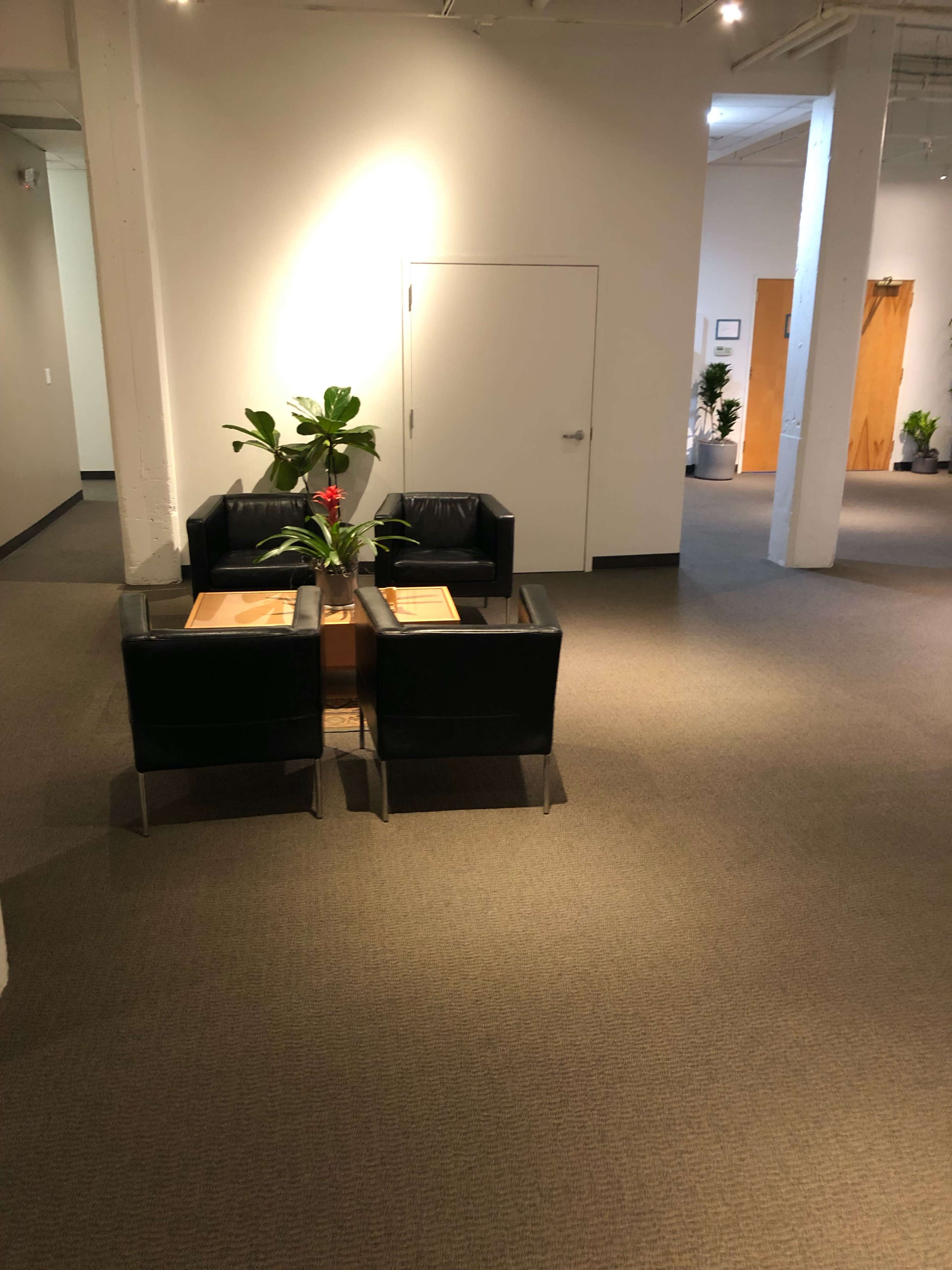 A spacious, well-lit office area with a small seating arrangement consisting of two black chairs and a coffee table near a potted plant.