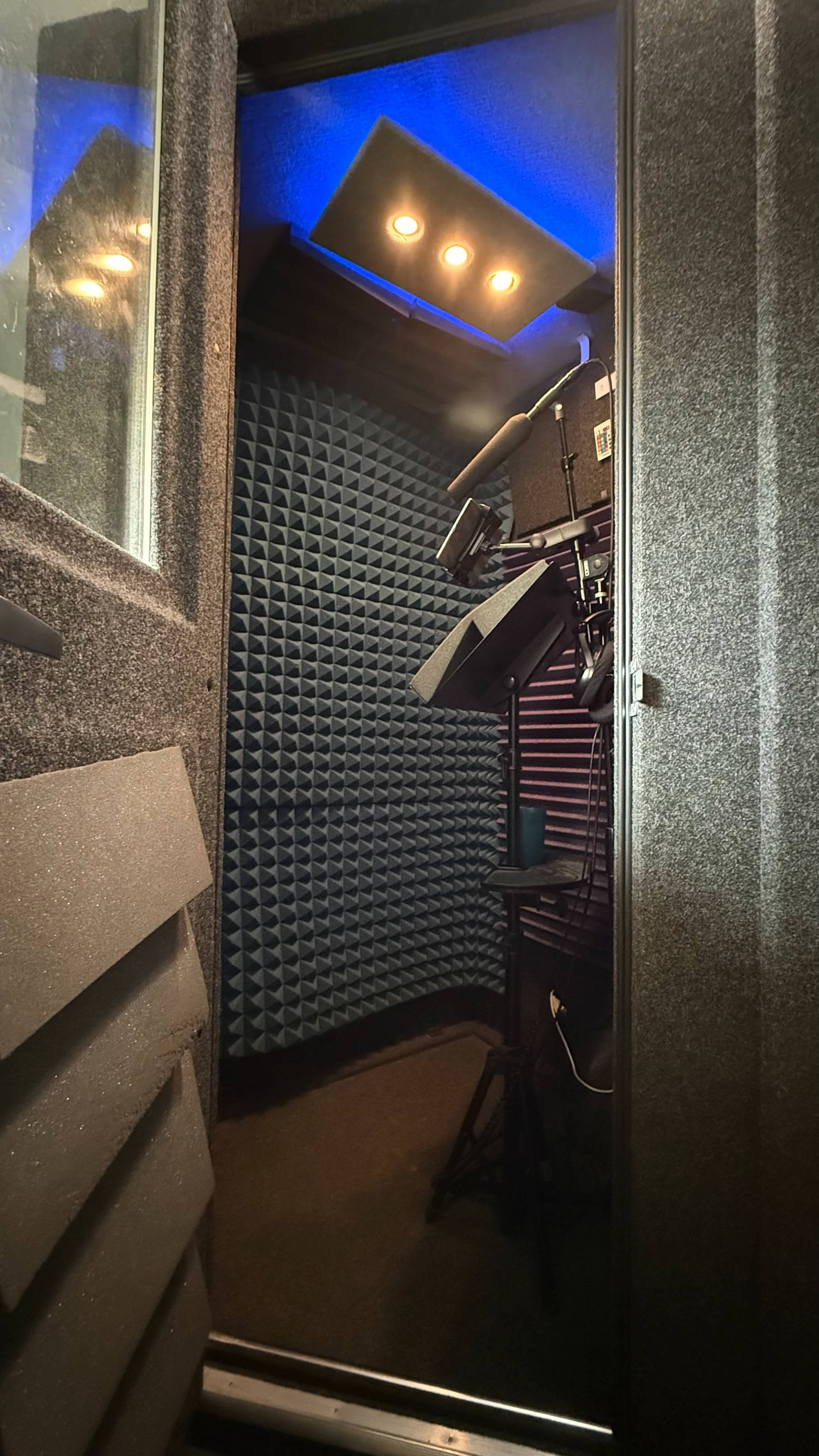 The image shows the interior of a soundproof recording booth with acoustic treatment and equipment set up.