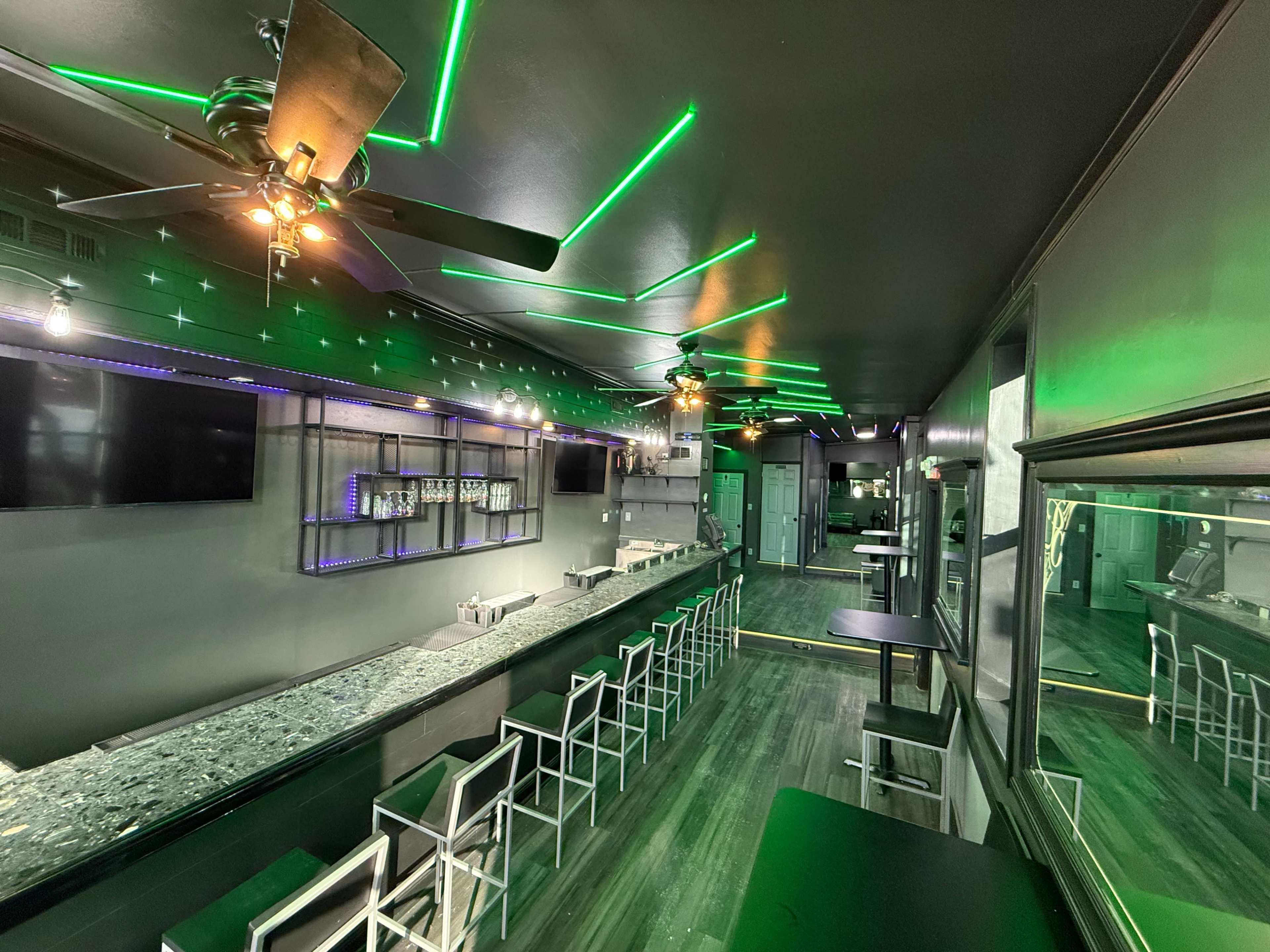 The image shows a modern bar interior with a long countertop, seating along one side, and green LED lights illuminating the ceiling.