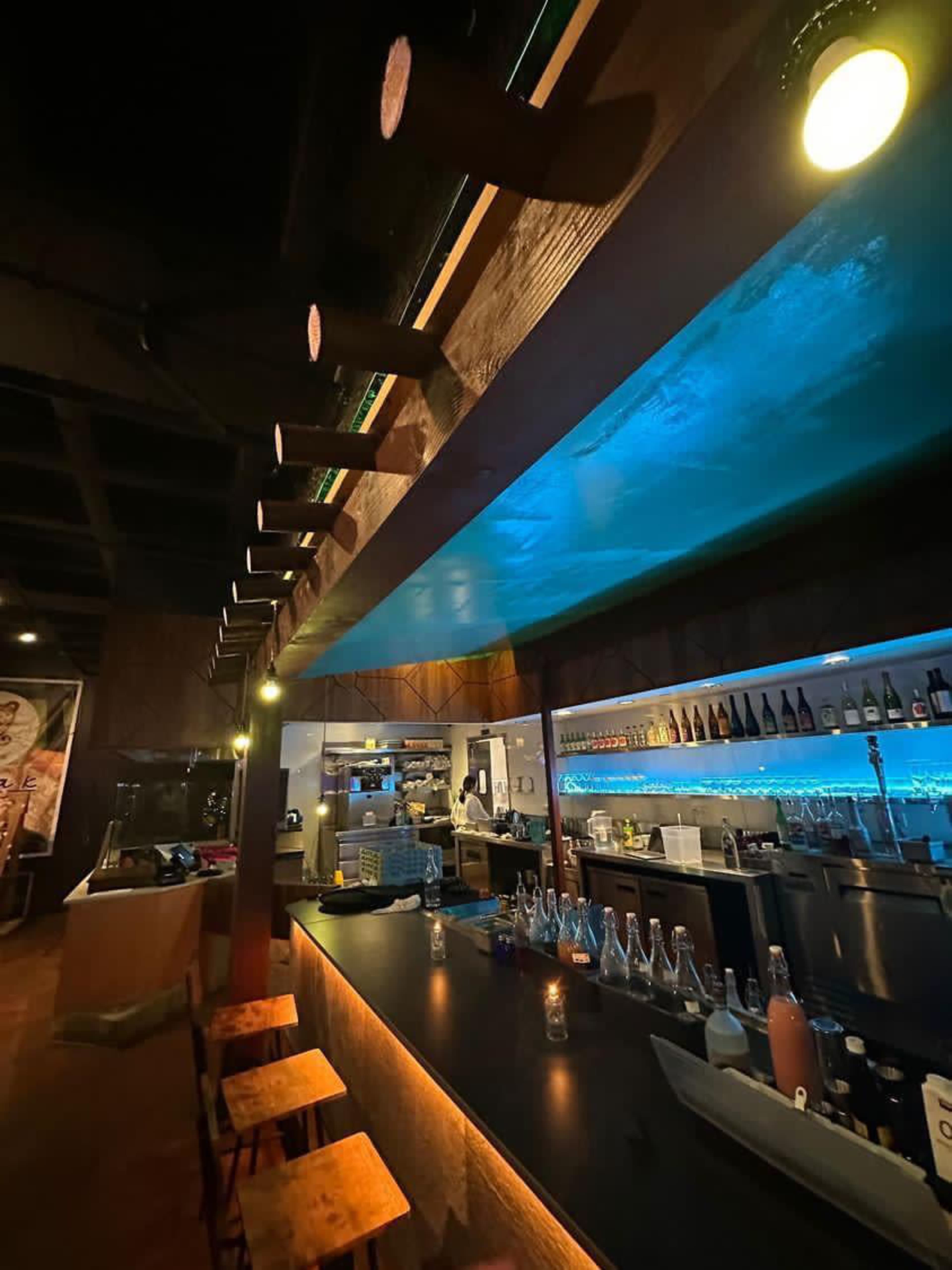 A modern bar area with wooden stools, a counter illuminated by blue and yellow lights, and shelves stocked with bottles.