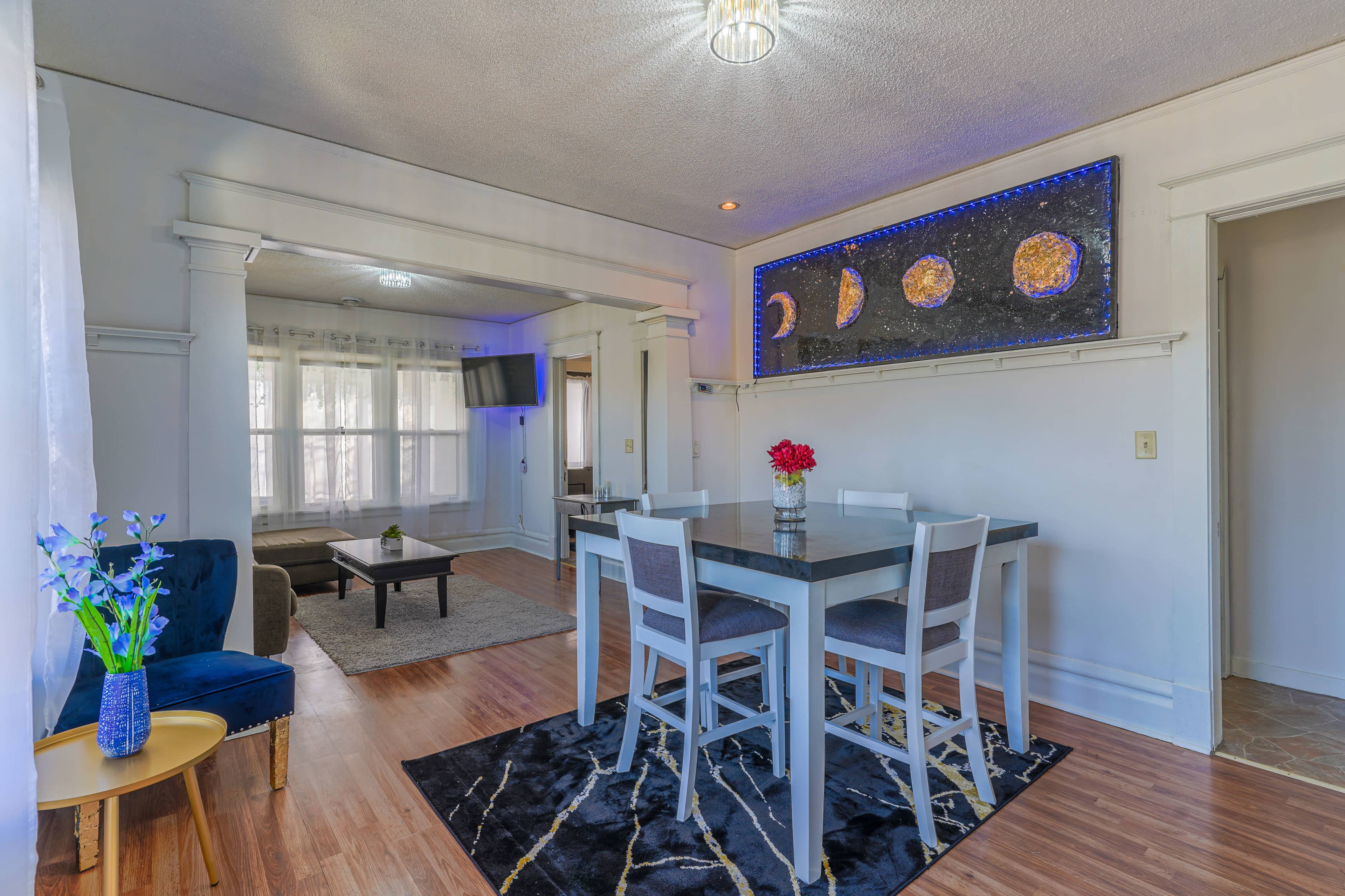 A dining area features a gray table surrounded by white chairs, with a floral centerpiece and a wall art piece illuminated by blue lighting.