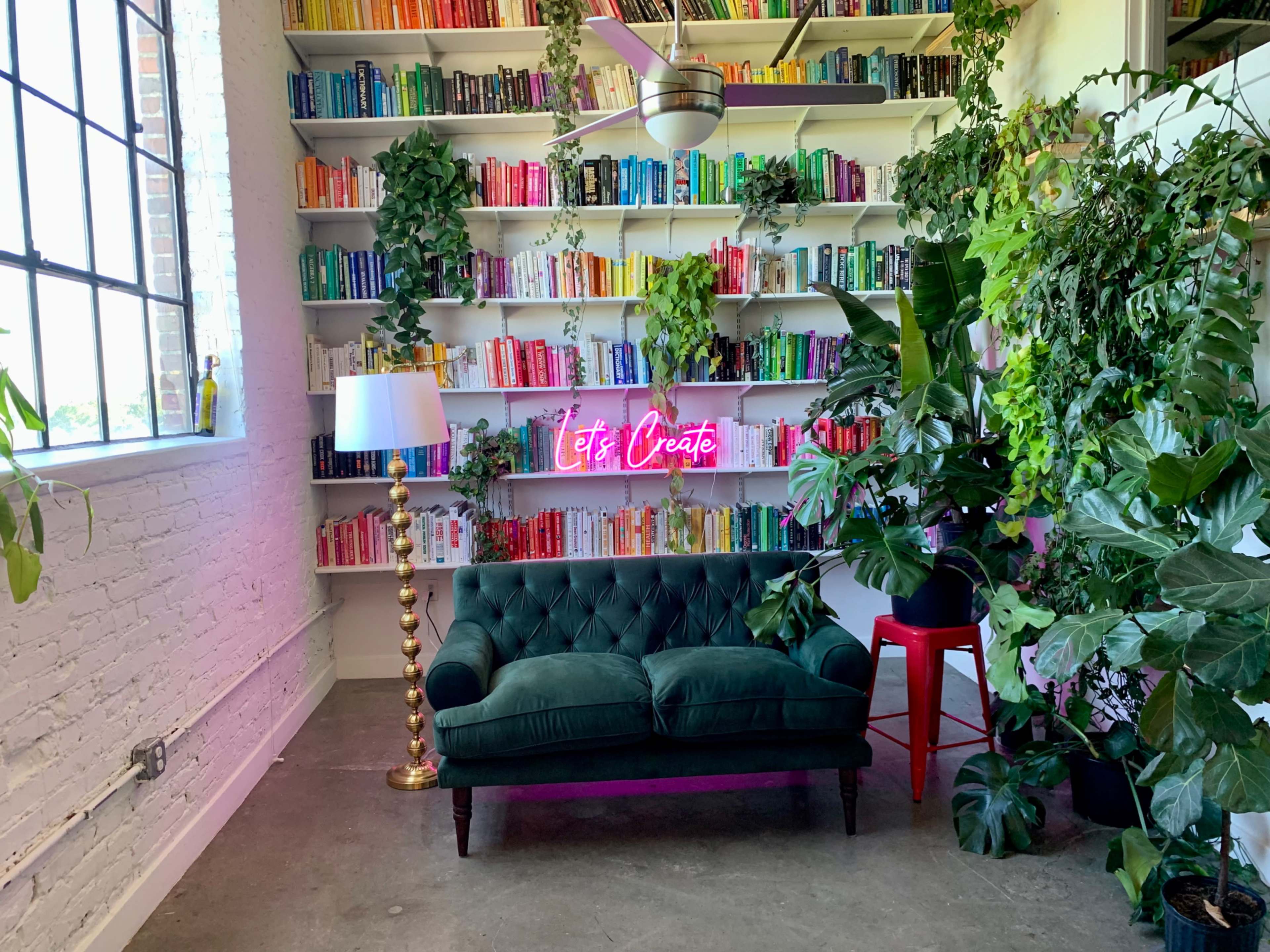 A green tufted sofa sits in front of a brightly colored bookshelf, surrounded by various plants and illuminated by a neon sign that reads "Let's Create."