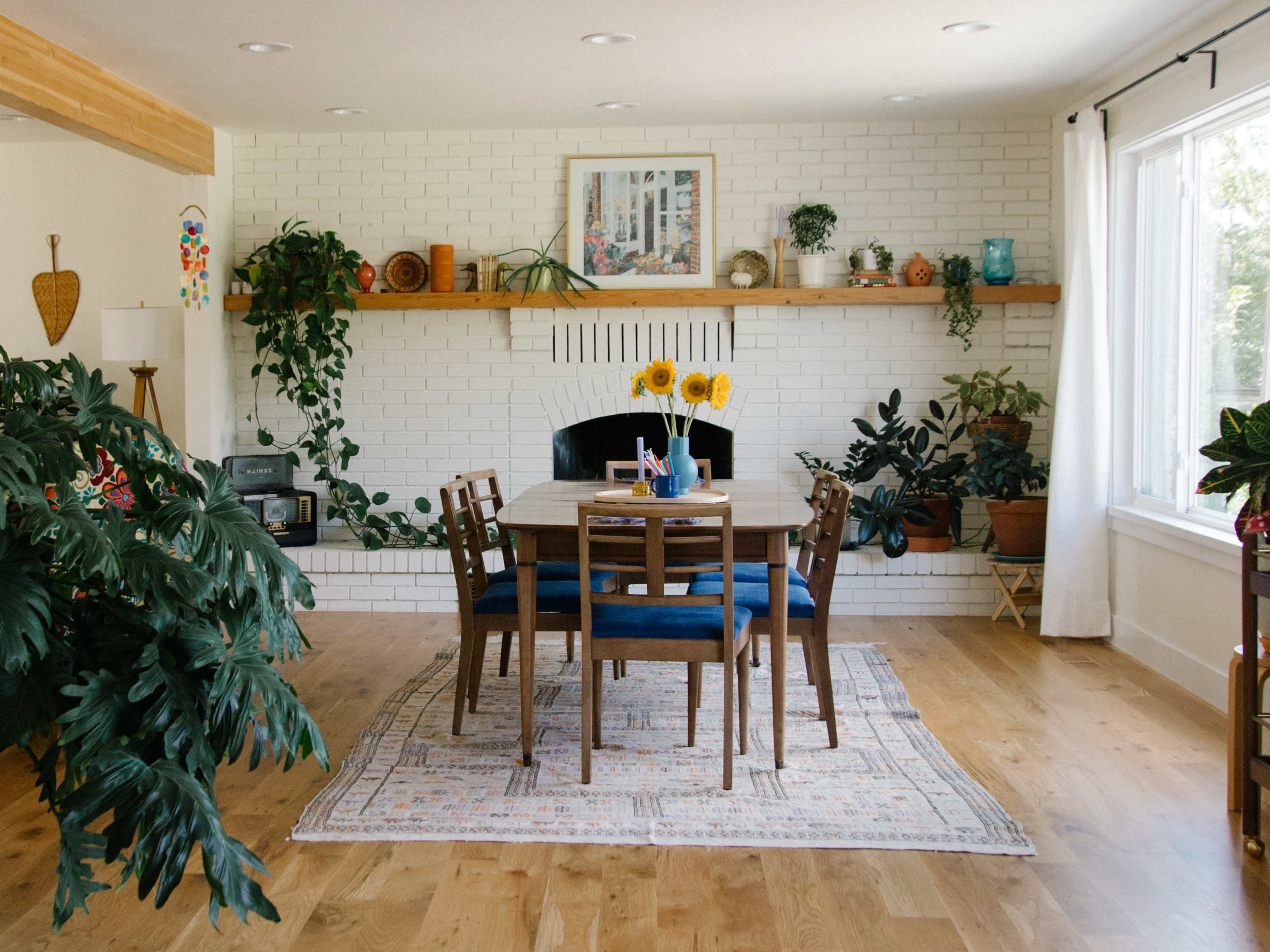 A dining area features a wooden table surrounded by chairs, with a fireplace and plants adorning the white brick wall.