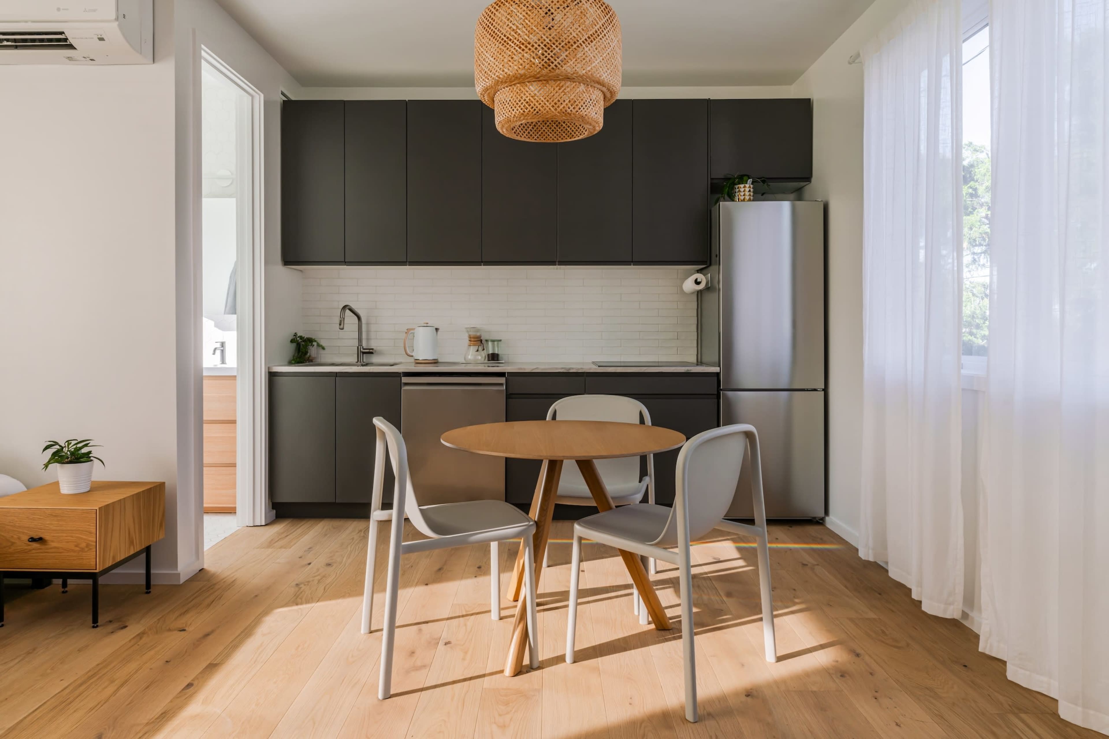 A modern kitchen features dark cabinetry, a stainless steel refrigerator, a round dining table with three chairs, and natural light streaming through sheer curtains.
