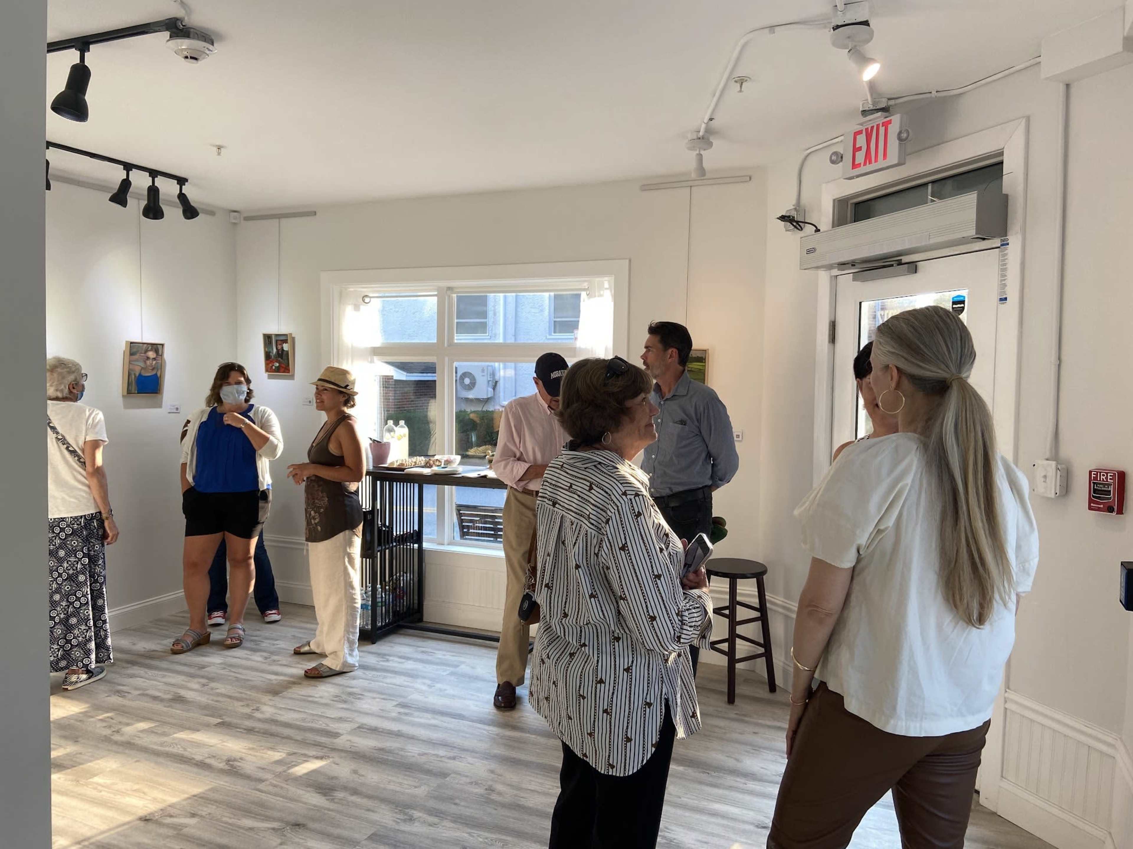 A group of people is gathered in a brightly lit art gallery, with several artworks displayed on the walls.