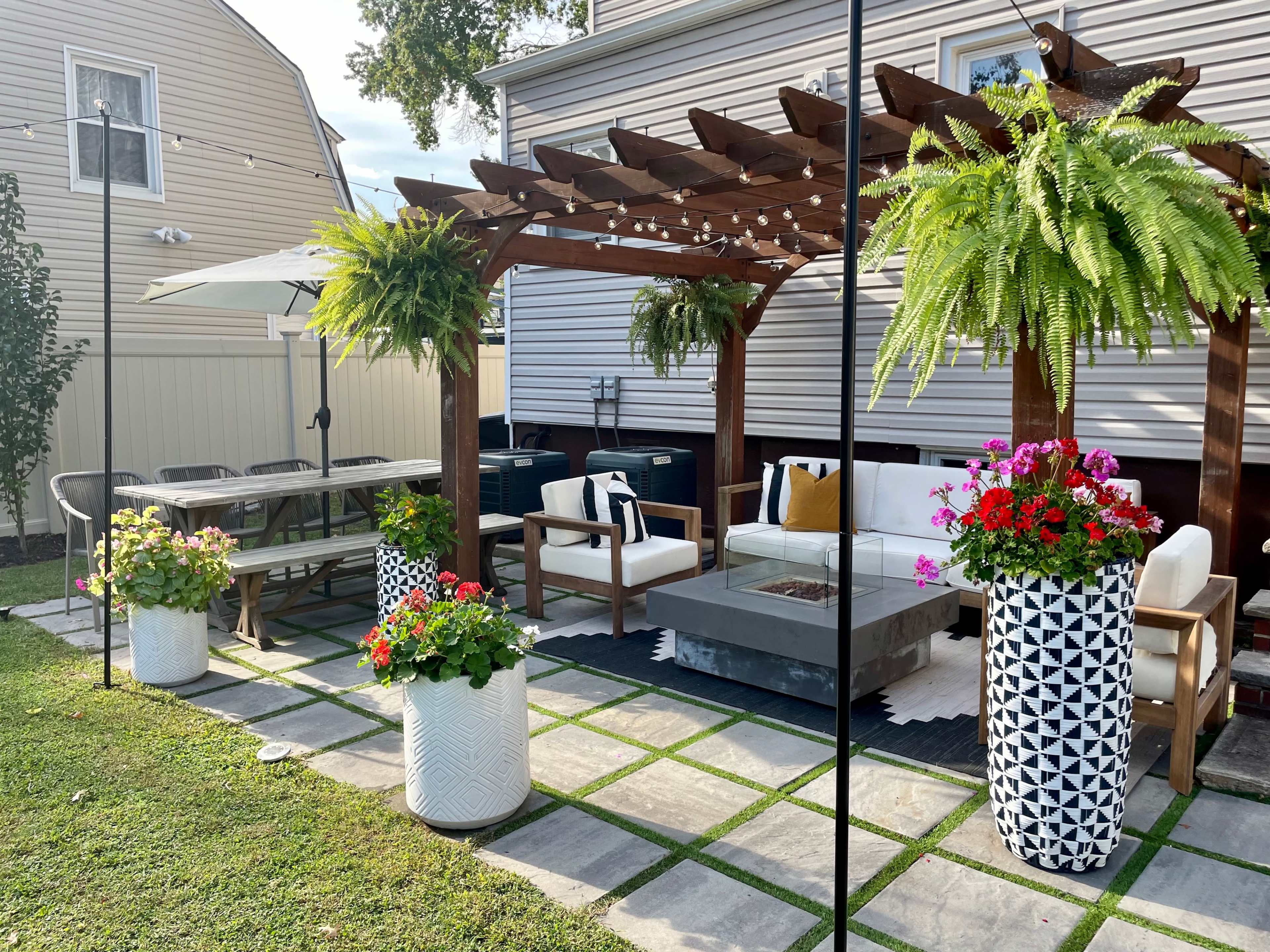 A backyard patio featuring a wooden pergola with hanging ferns, a seating area with a gray coffee table and white couch, and large flower planters filled with colorful plants.