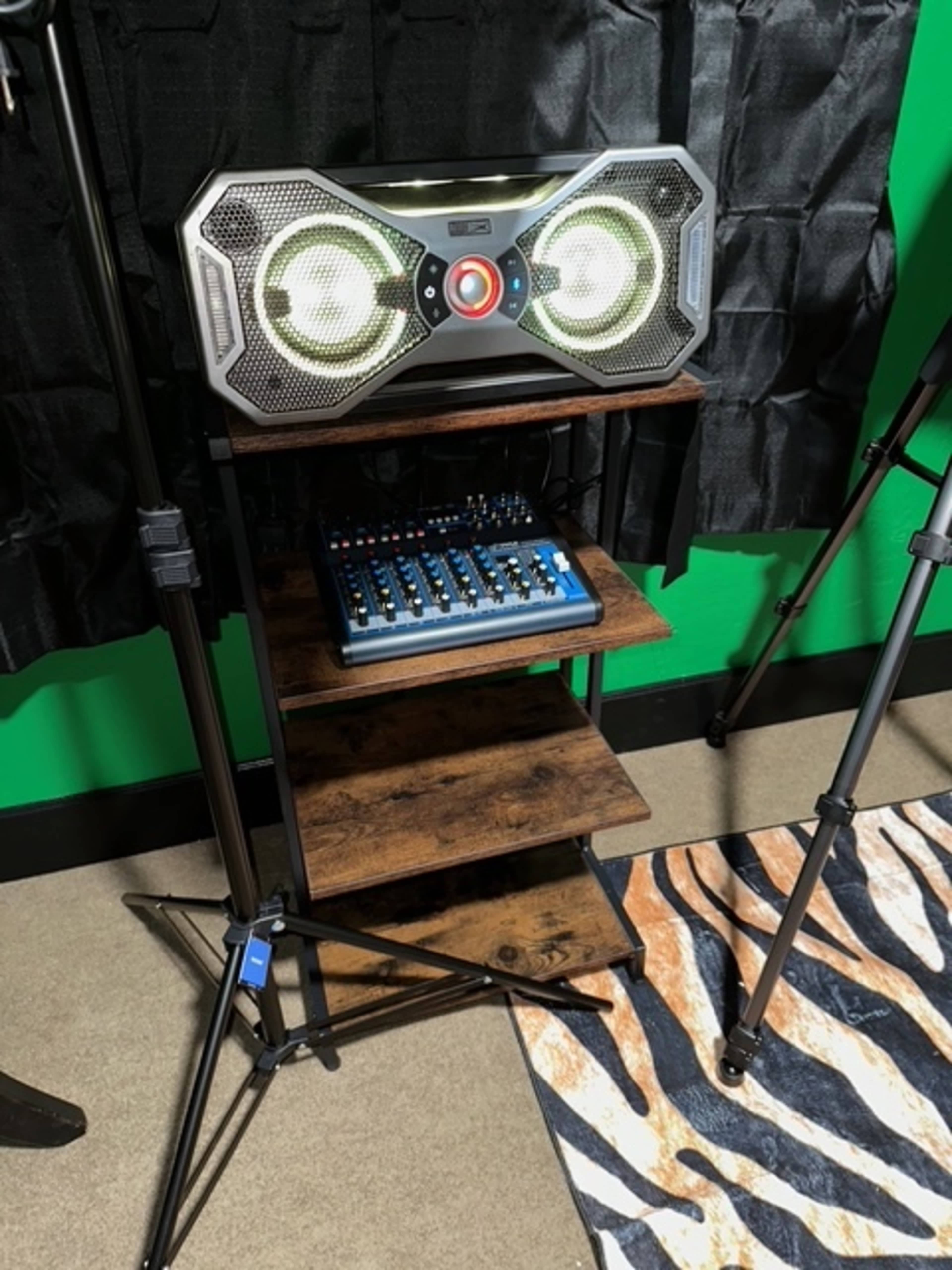 The image shows a wooden shelf stand with a large stereo speaker on top and a blue audio mixer below, set against a green wall and black curtains.