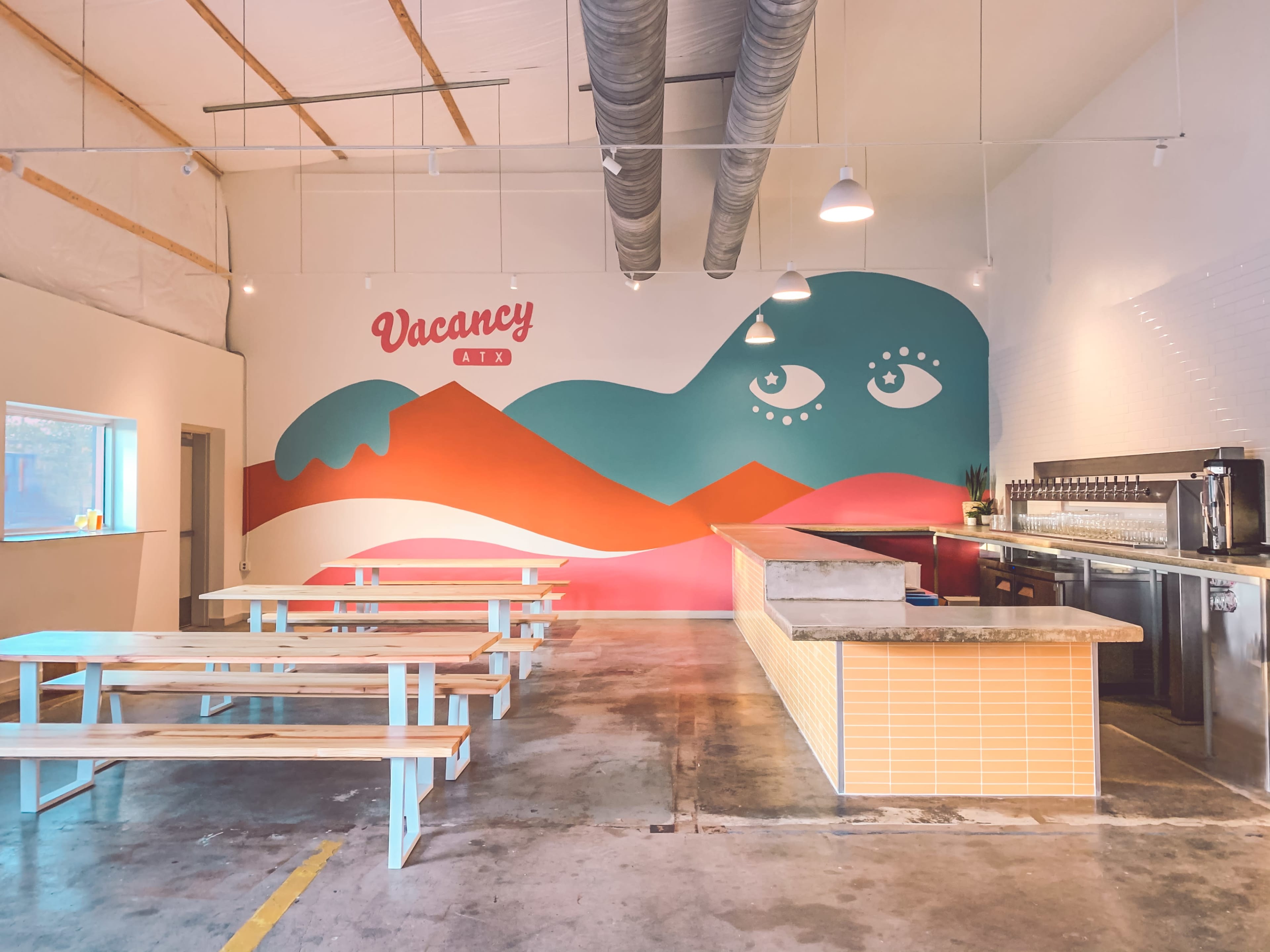 The image shows a spacious cafe interior with a large mural featuring abstract shapes and eyes on one wall, complemented by wooden tables aligned in the center and a serving area at the back.