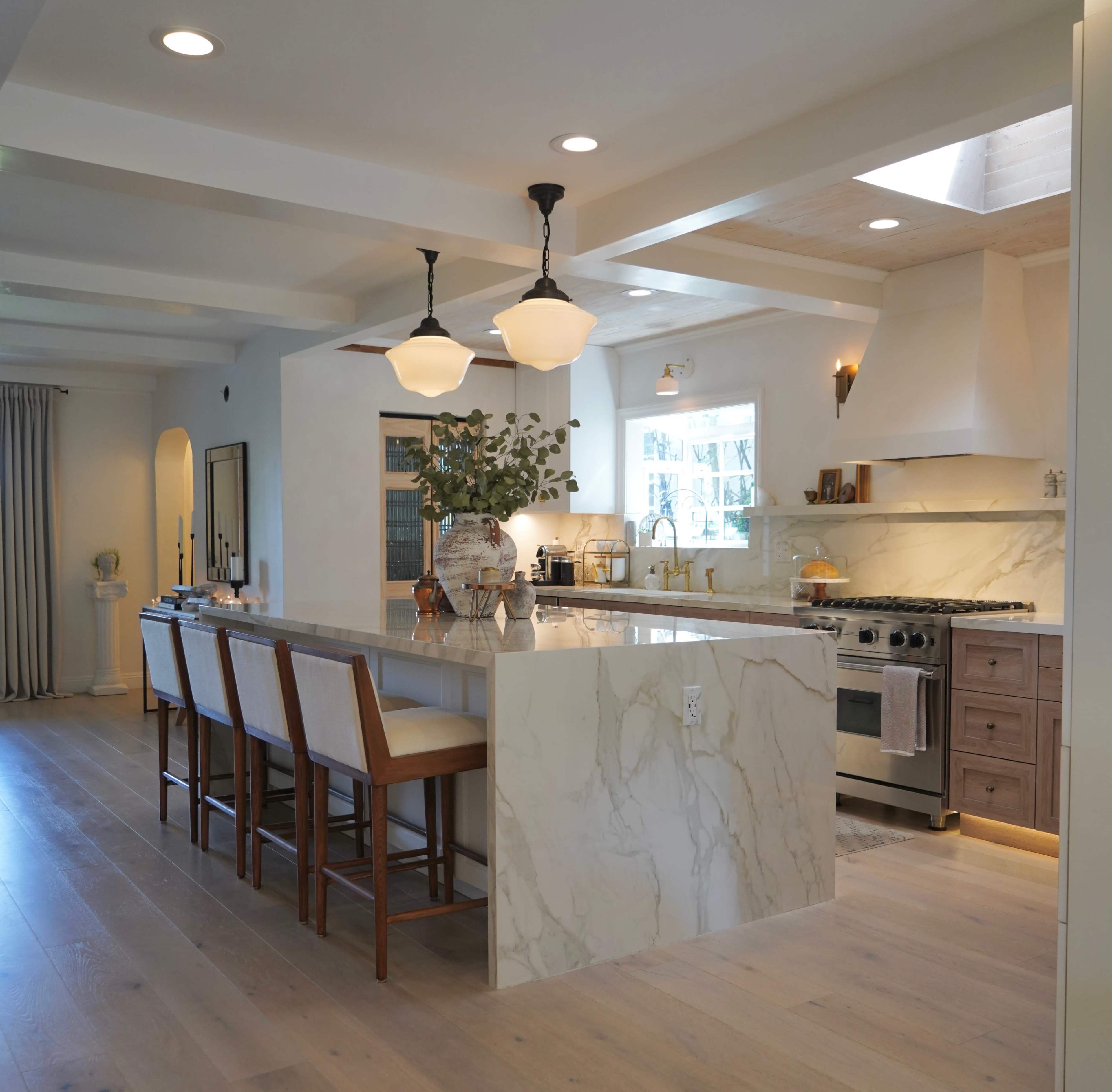 The image shows a modern kitchen with a spacious island featuring marble countertops, pendant lighting, and a stove with an exhaust hood, surrounded by wooden barstools.