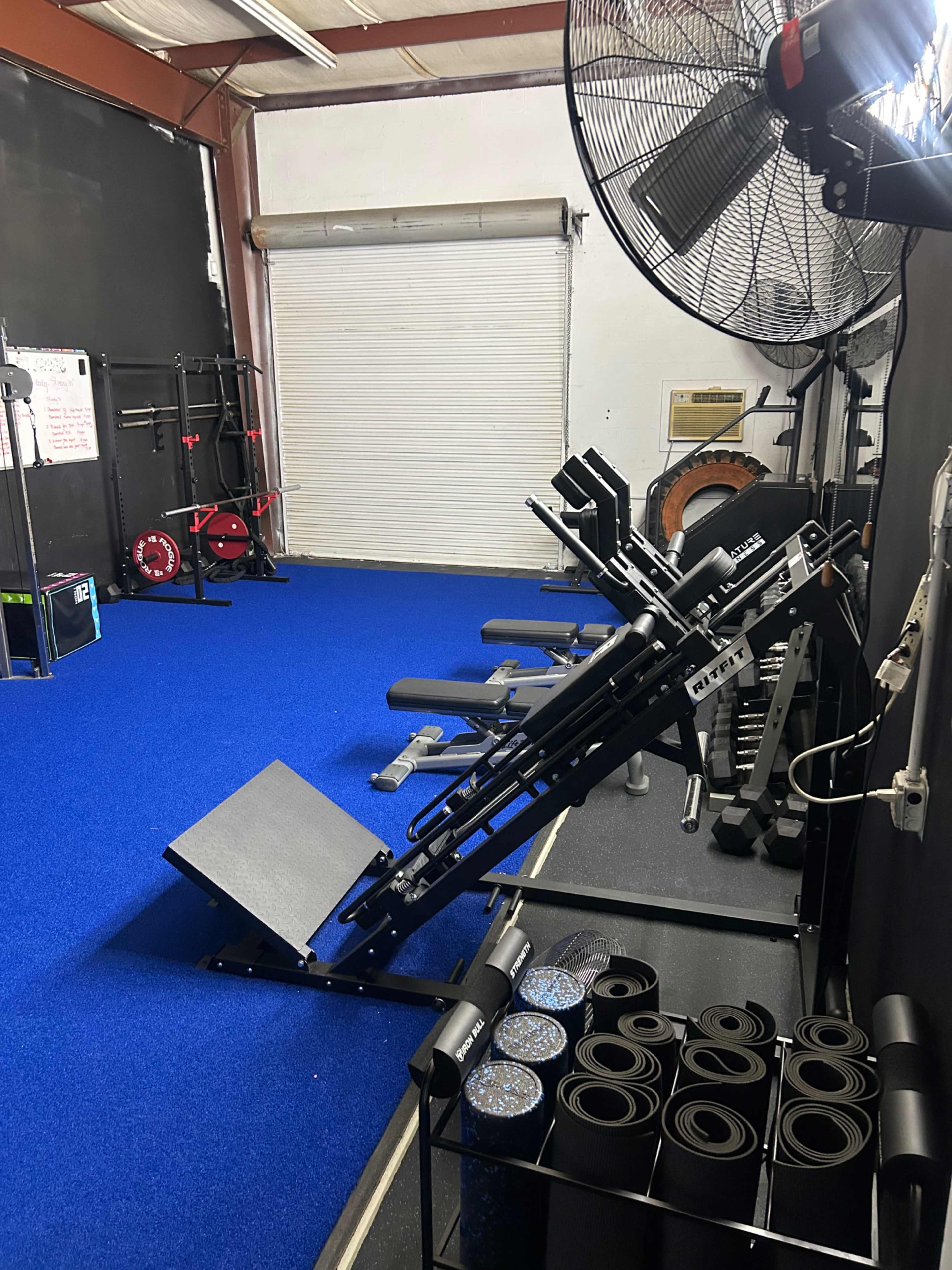 The image shows a gym interior with various fitness equipment, including benches, weights, and exercise mats, against a backdrop of blue flooring and a fan.