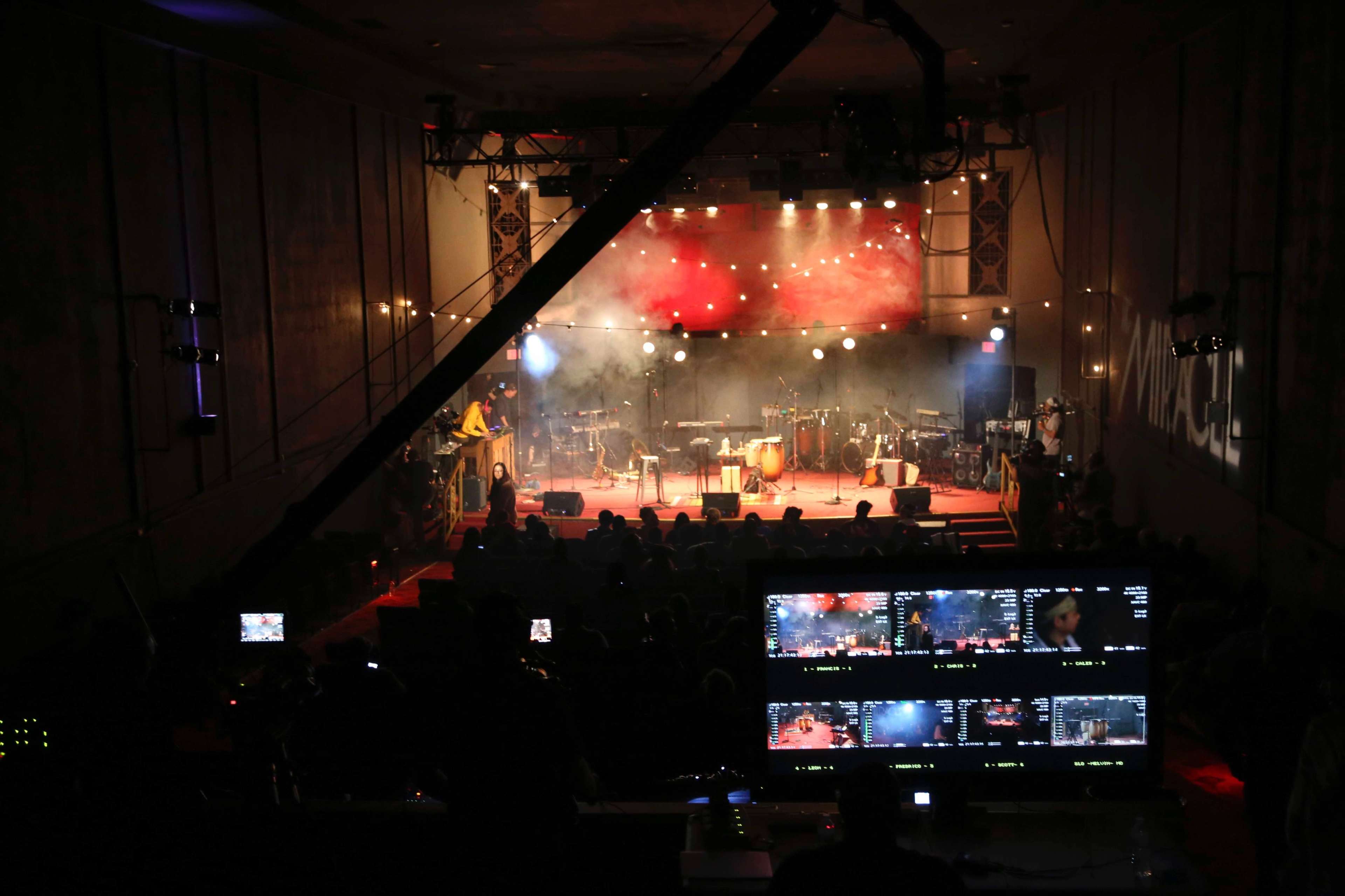 A dimly lit auditorium set up for a live performance, featuring a stage with musical instruments and a control station with multiple video monitors in the foreground.