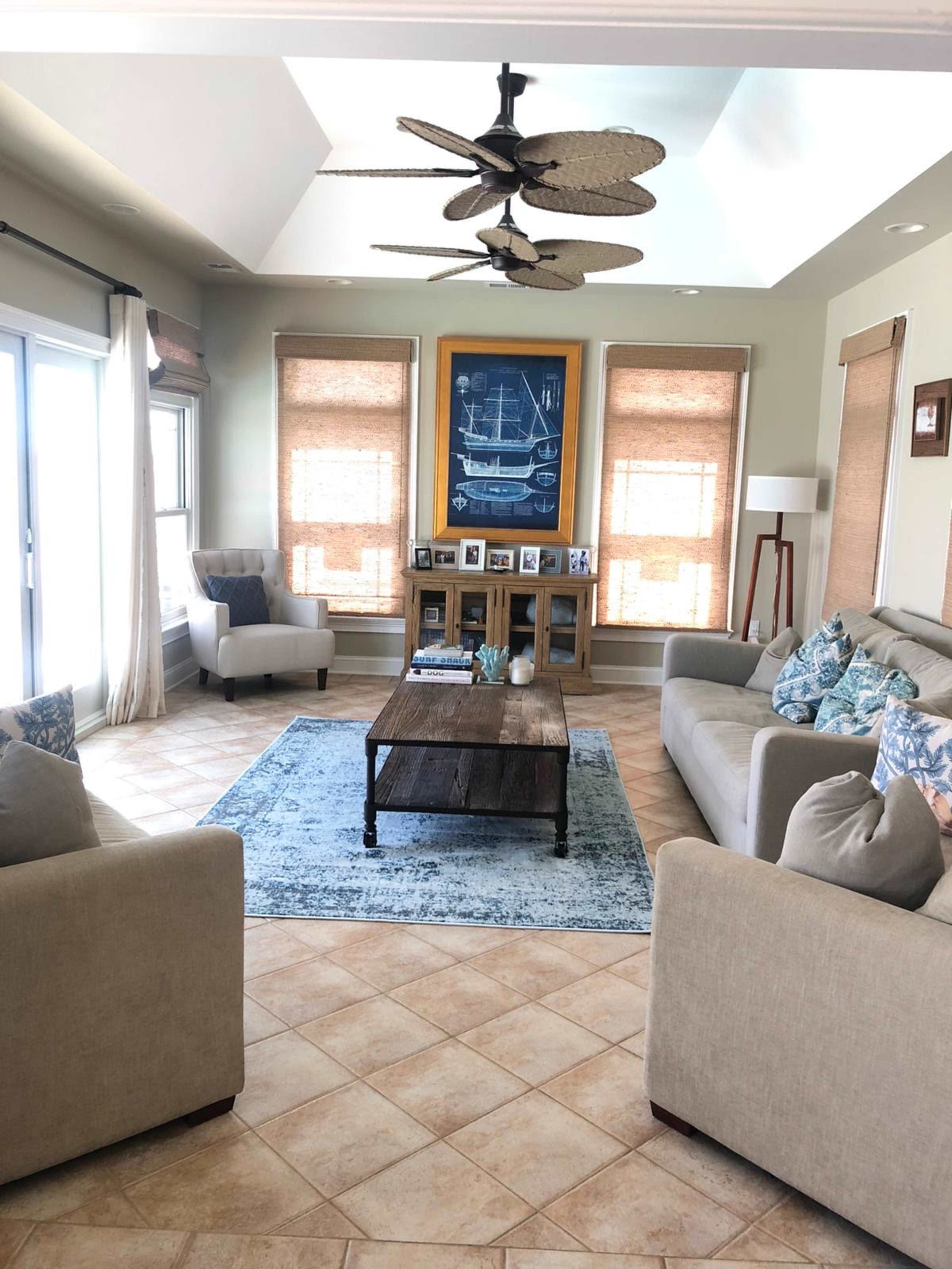 A light-filled living room with two sofas, a coffee table, and a decorative area rug, surrounded by large windows and framed photographs on a shelf.
